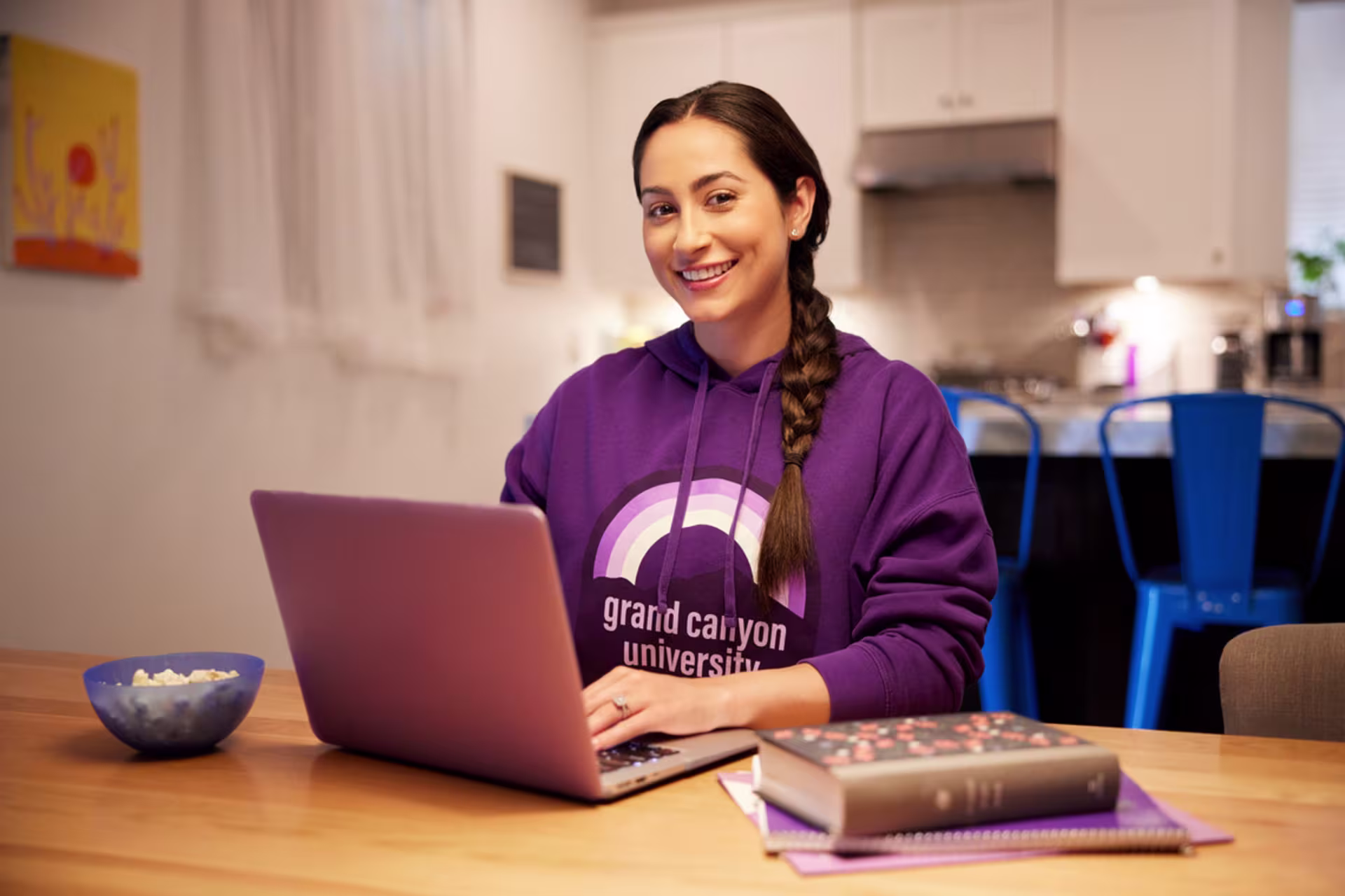 A smiling female student wearing a Grand Canyon University hoodie works on a laptop at a kitchen table with books nearby.