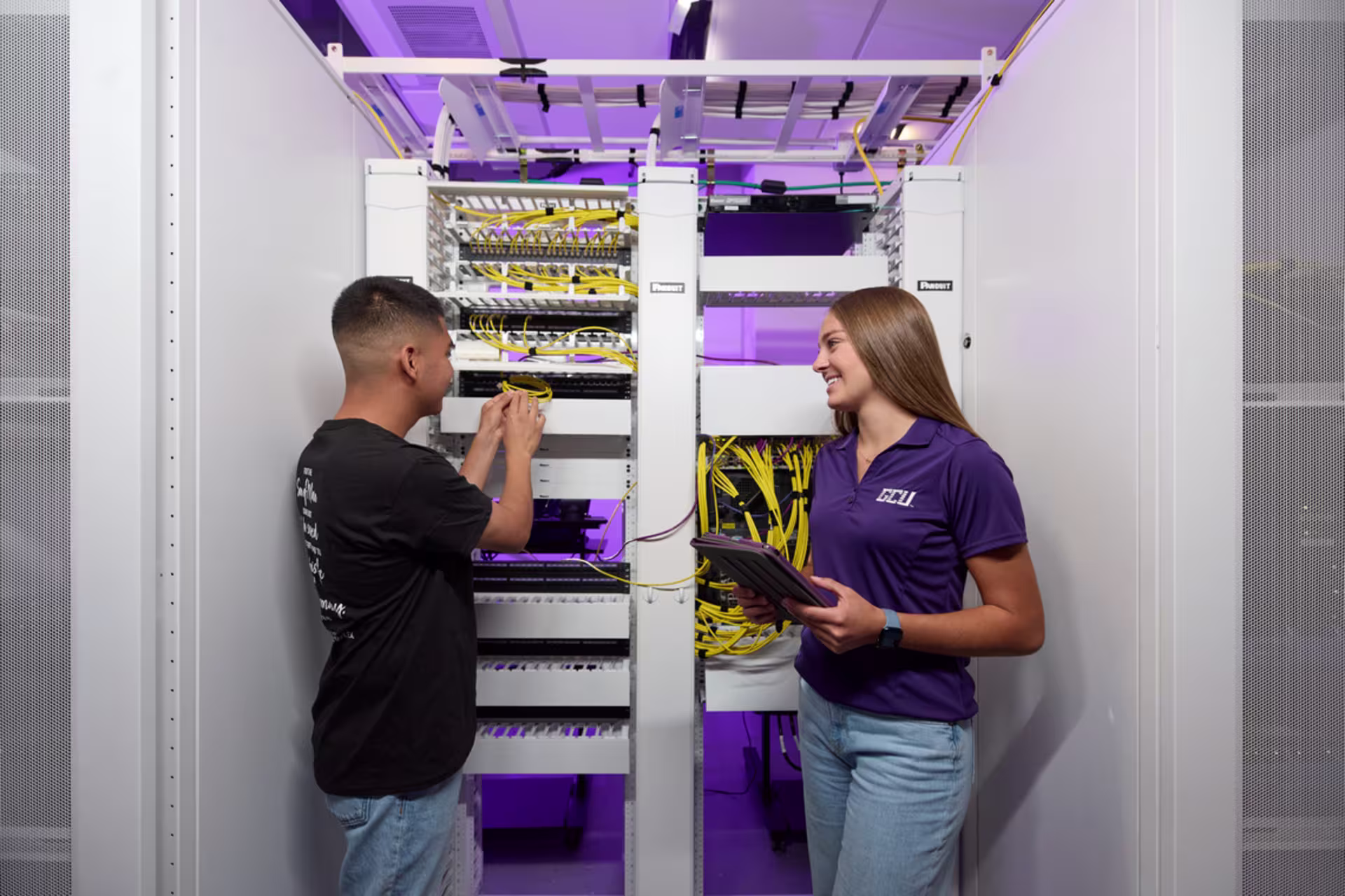 Two GCU students examine networking equipment in an open server rack, with visible cables, switches and GCU branded shirts in a technology lab.