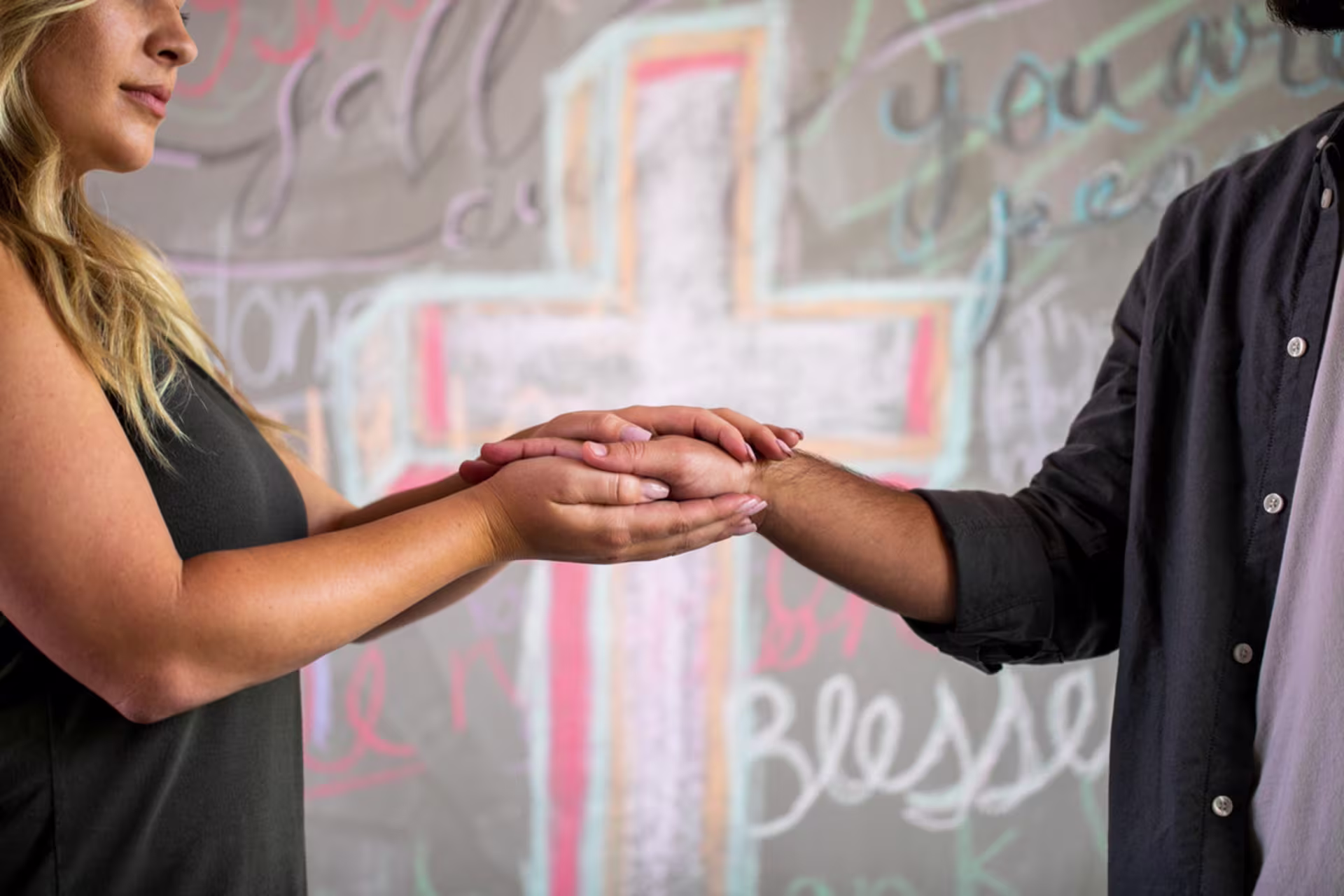 Two people gently hold hands in front of a wall with a softly colored cross and handwritten words in the background, suggesting a moment of support or prayer.
