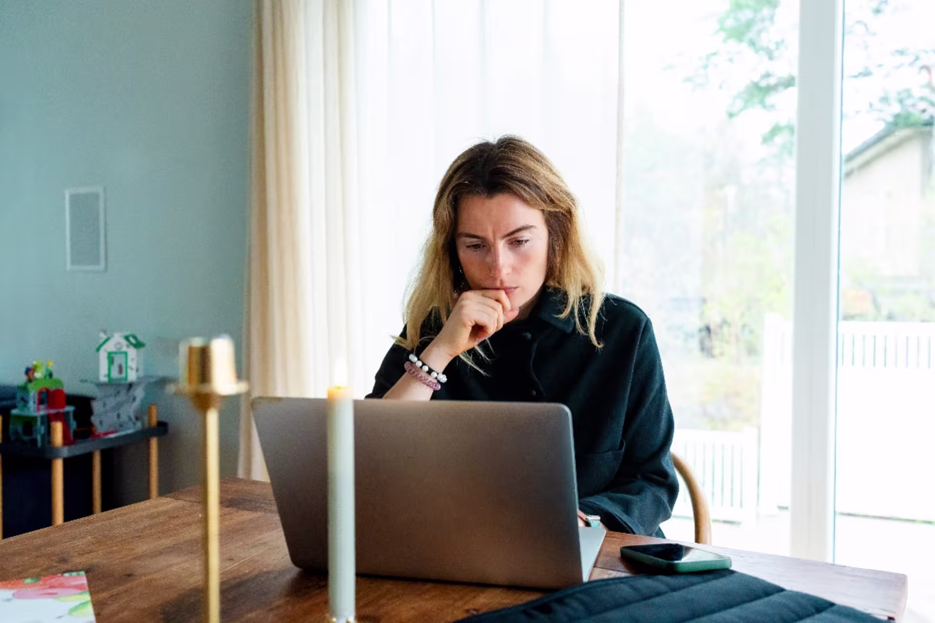 A woman sits at a wooden table working on a laptop, resting her chin on her hand as she looks thoughtfully at the screen.
