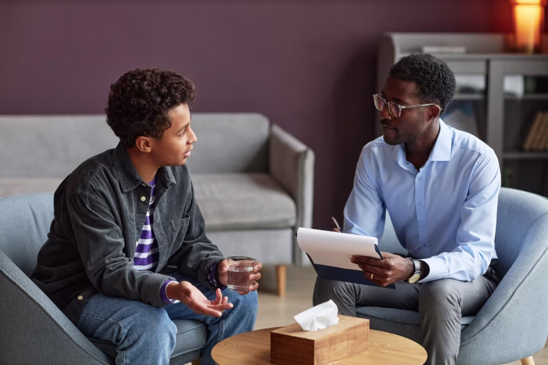 A psychologist listens and takes notes while speaking with a young client during a counseling session in an office setting.