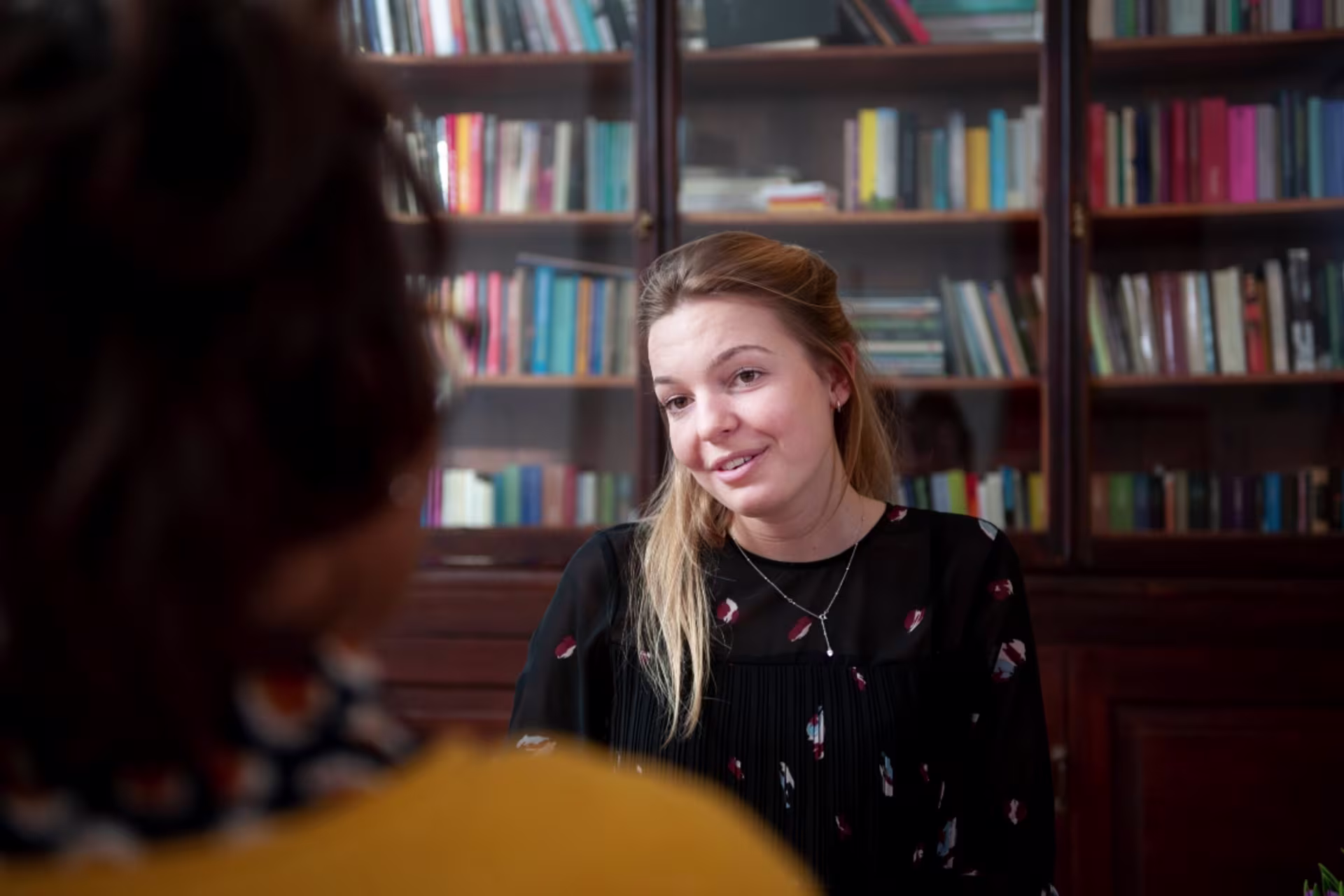 A psychologist listens attentively while speaking with a client during a counseling session in an office with bookshelves.