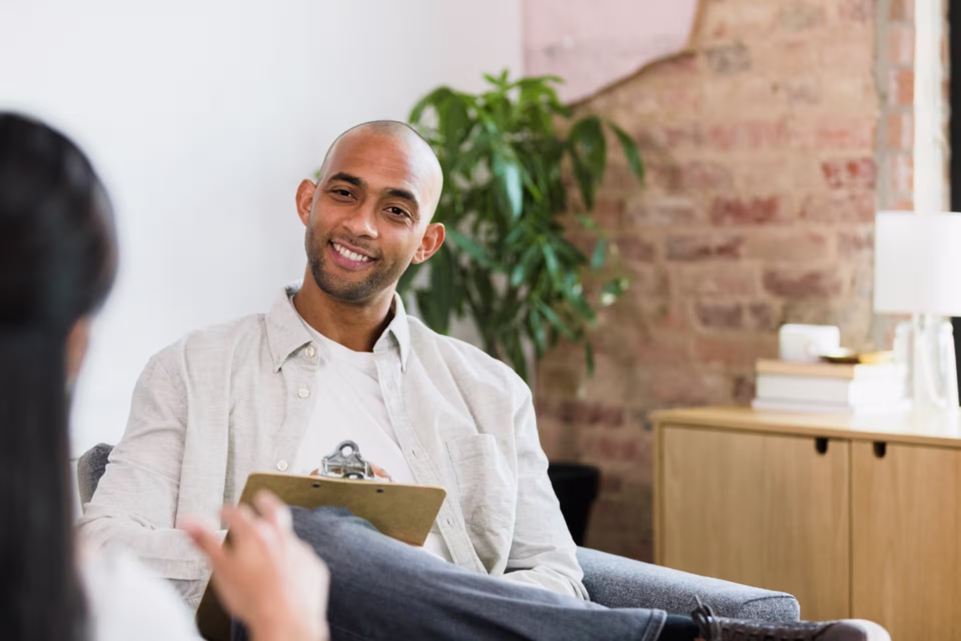 A psychologist smiles while holding a clipboard and speaking with a client during a counseling session in an office.
