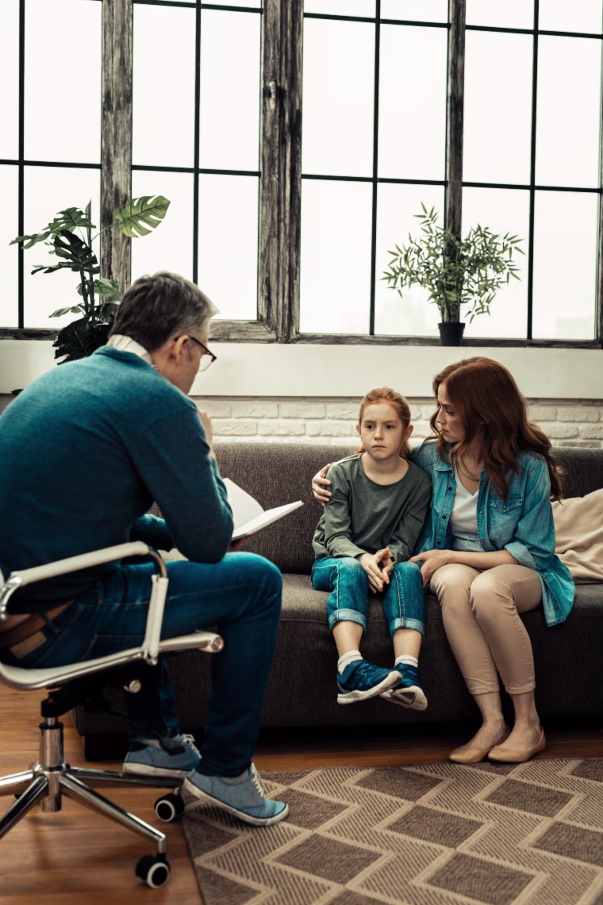Child and parent sitting on a couch during a counseling session while a therapist takes notes nearby.