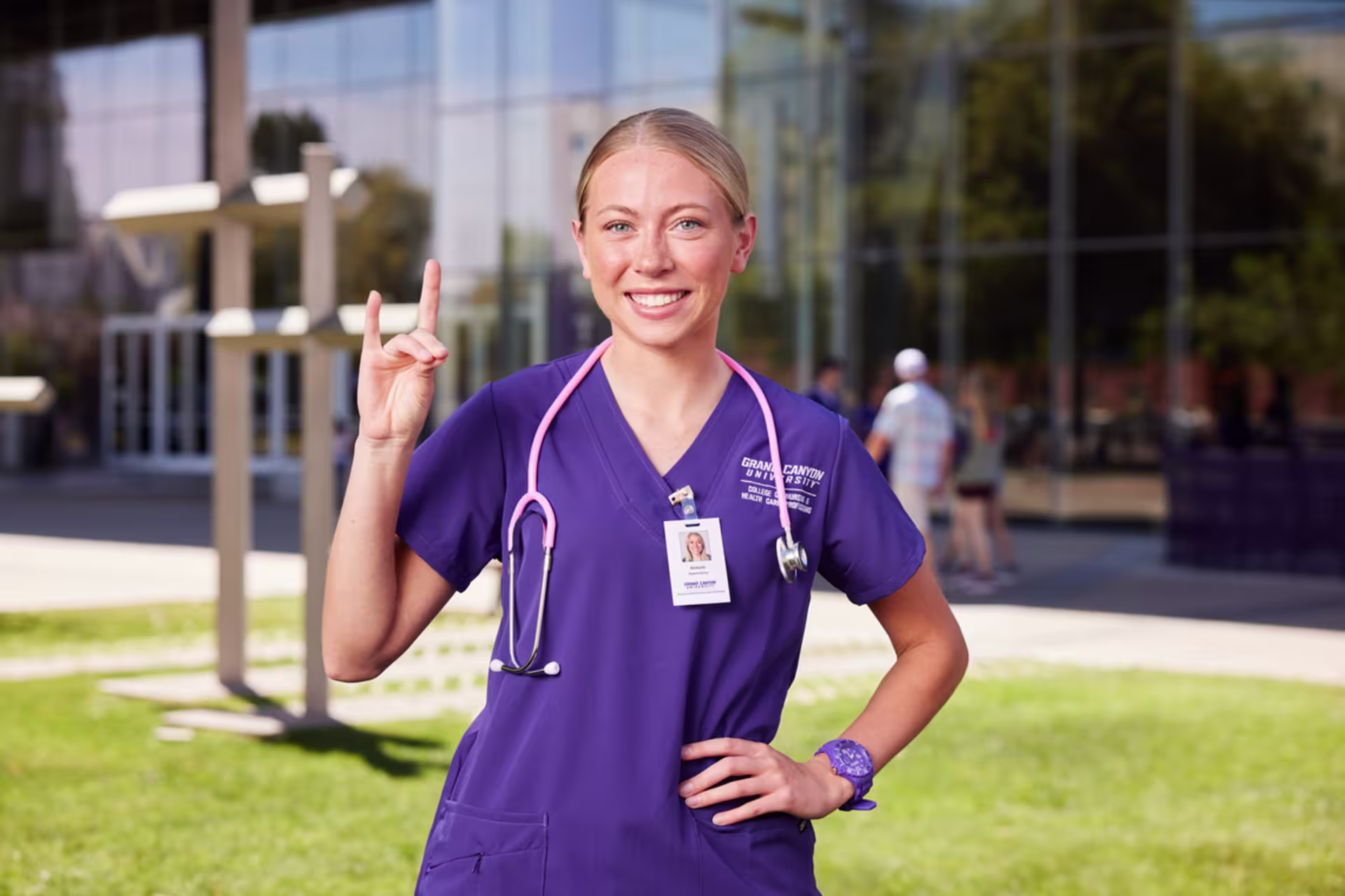 A female APRN student stands outside wearing purple scrubs with a stethoscope around her neck while holding up the Lopes hands signal.