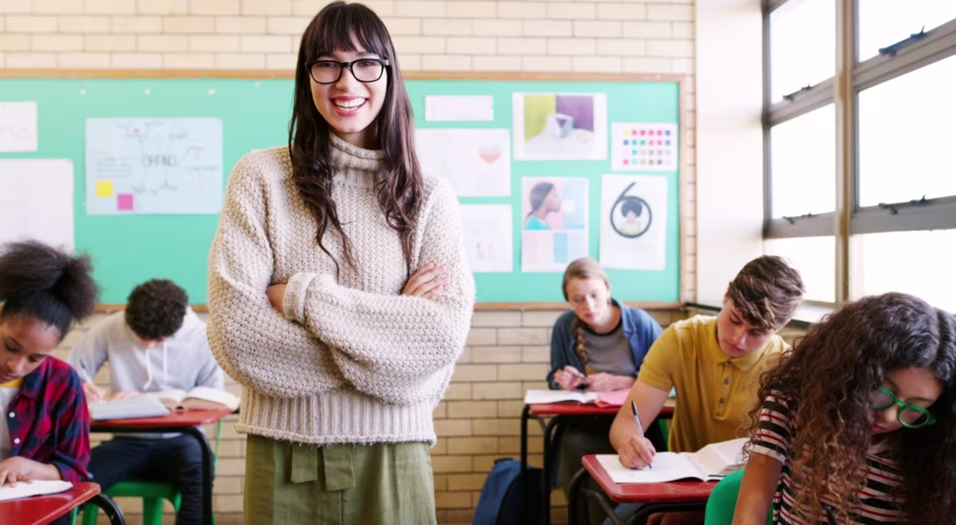 Female teacher stands with arms crossed in a classroom, with students working at desks in the background.