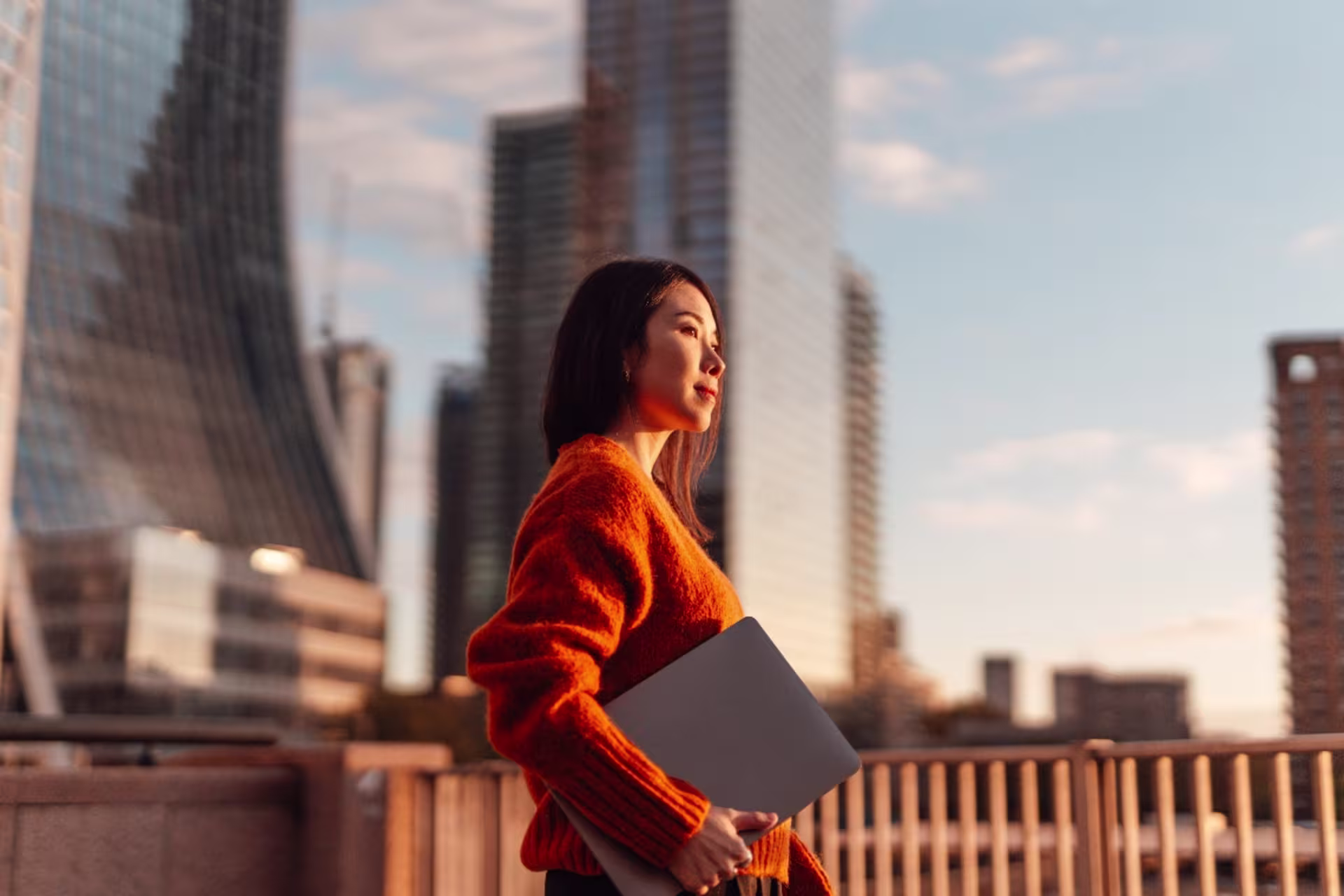 Woman holding a laptop on a city balcony at sunset, overlooking a modern skyline.
