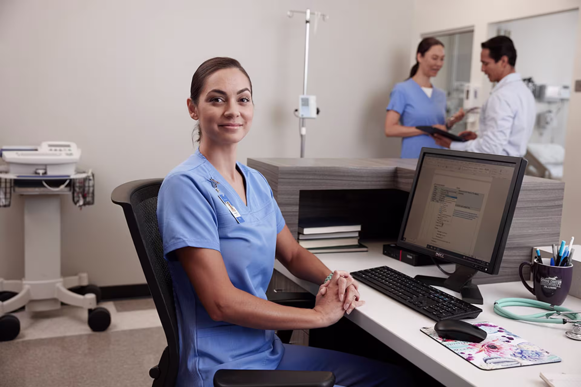 Female nurse sitting at a computer smiling with her hands folded.