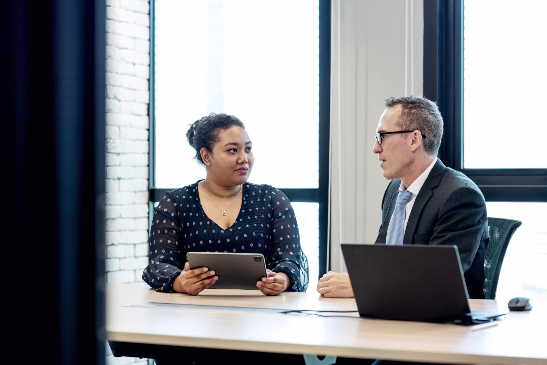 Two people sit at a table in a modern office, with one person holding a tablet while the other has an open laptop, appearing to discuss work.