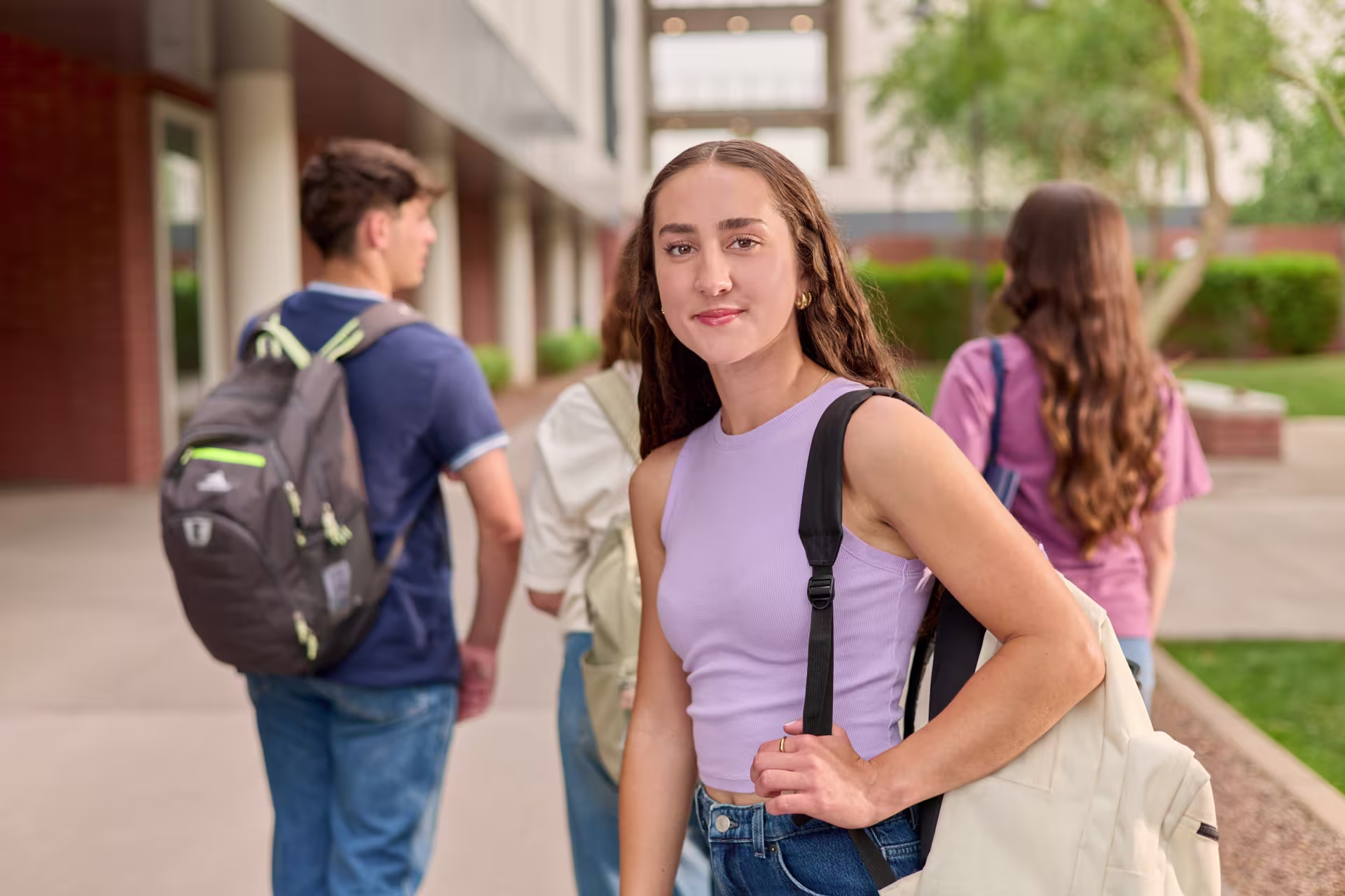 Female undergraduate student in a purple tank top walking on campus with a backpack with students in the background.