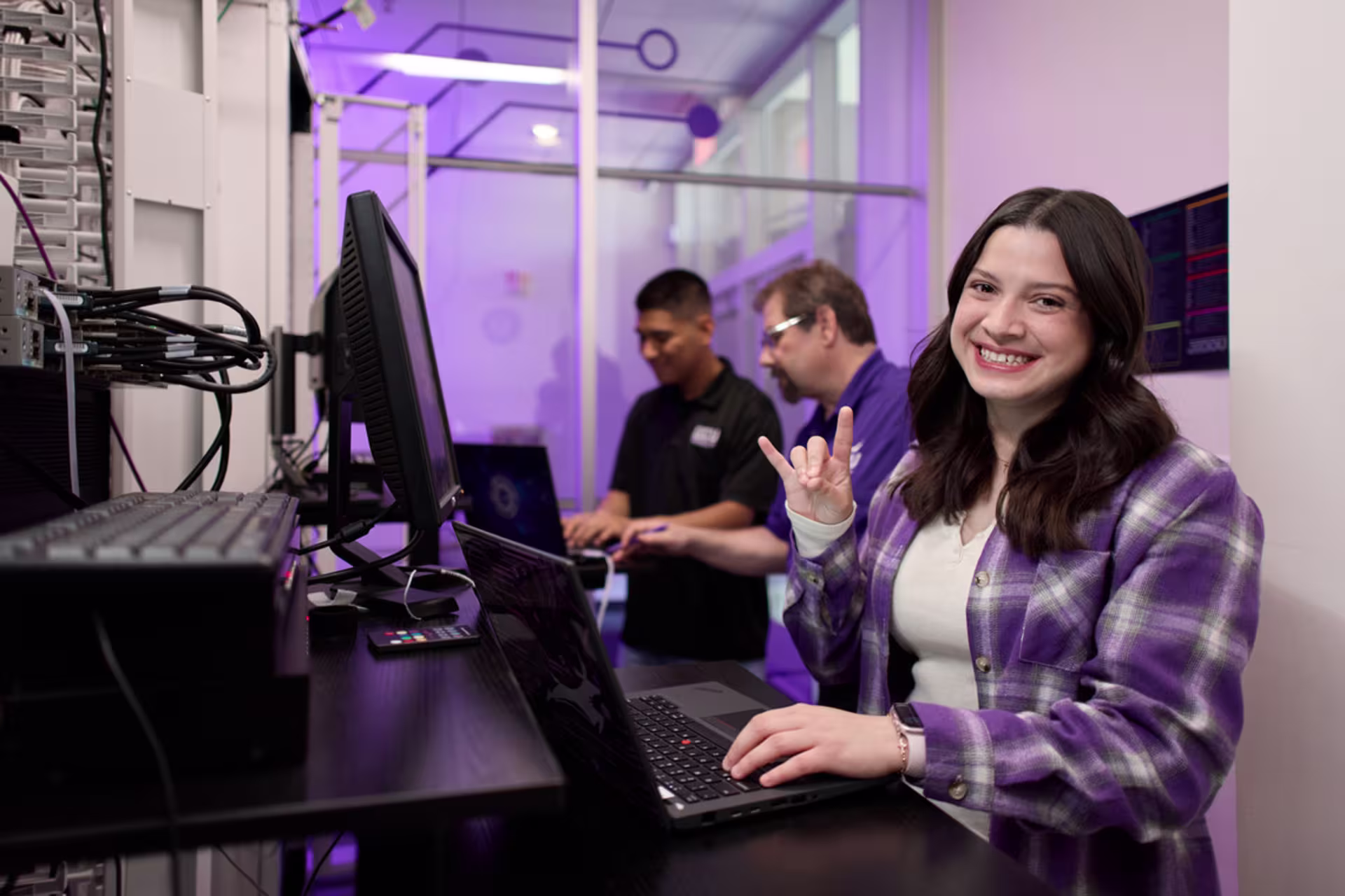 Young woman in a purple plaid shirt in a server room smiling at the camera while holding a laptop and making Lopes Up hand sign as a cybersecurity professor helps another student behind her, surrounded by racks of networking equipment and cables.