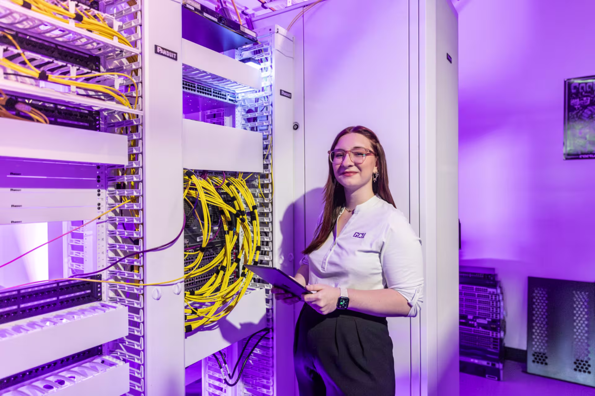 Female computer networking student stands beside a server rack filled with organized yellow Ethernet cables and switches, holding a tablet while monitoring infrastructure.
