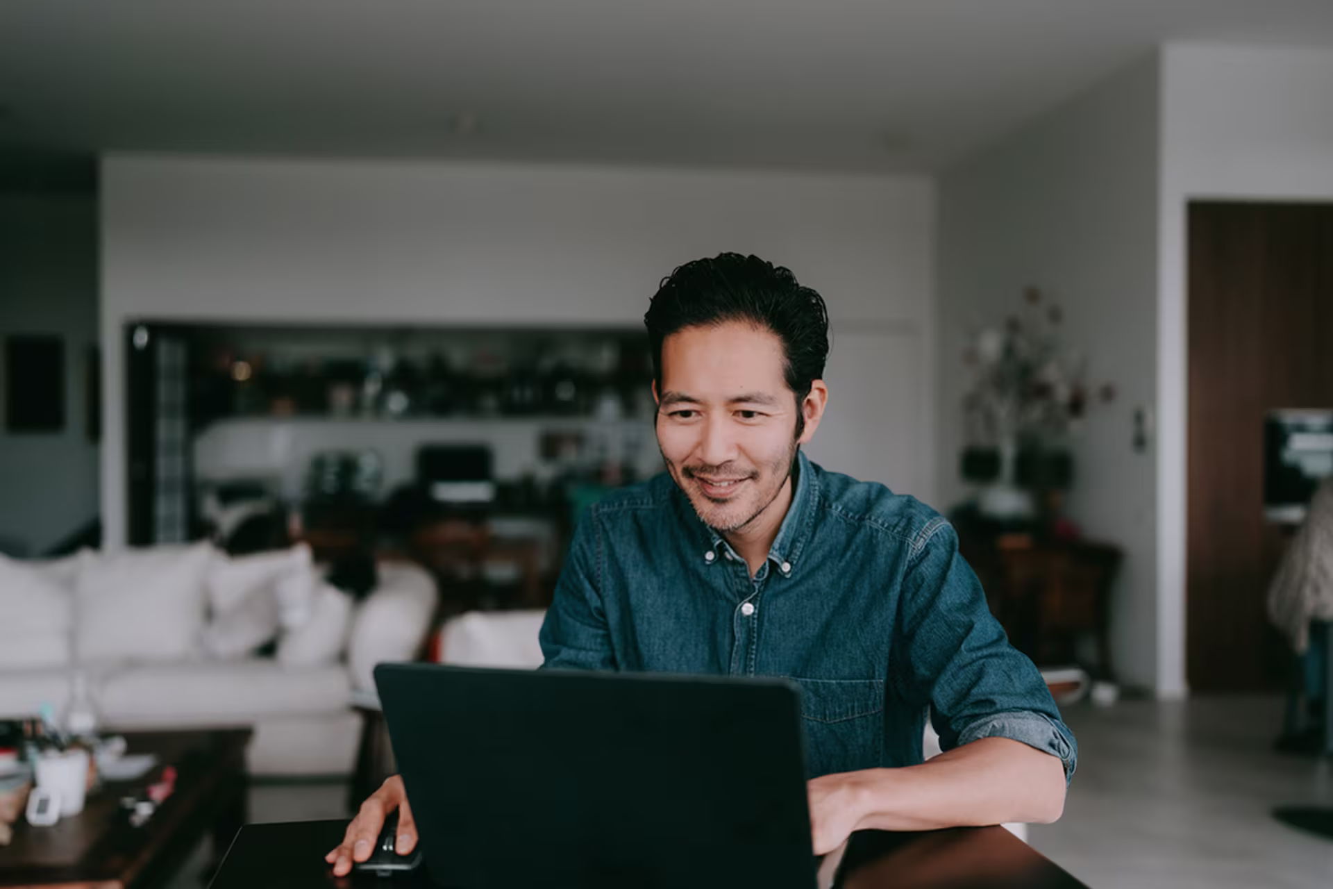 Man smiling while working on a laptop at home in a modern living room setting.