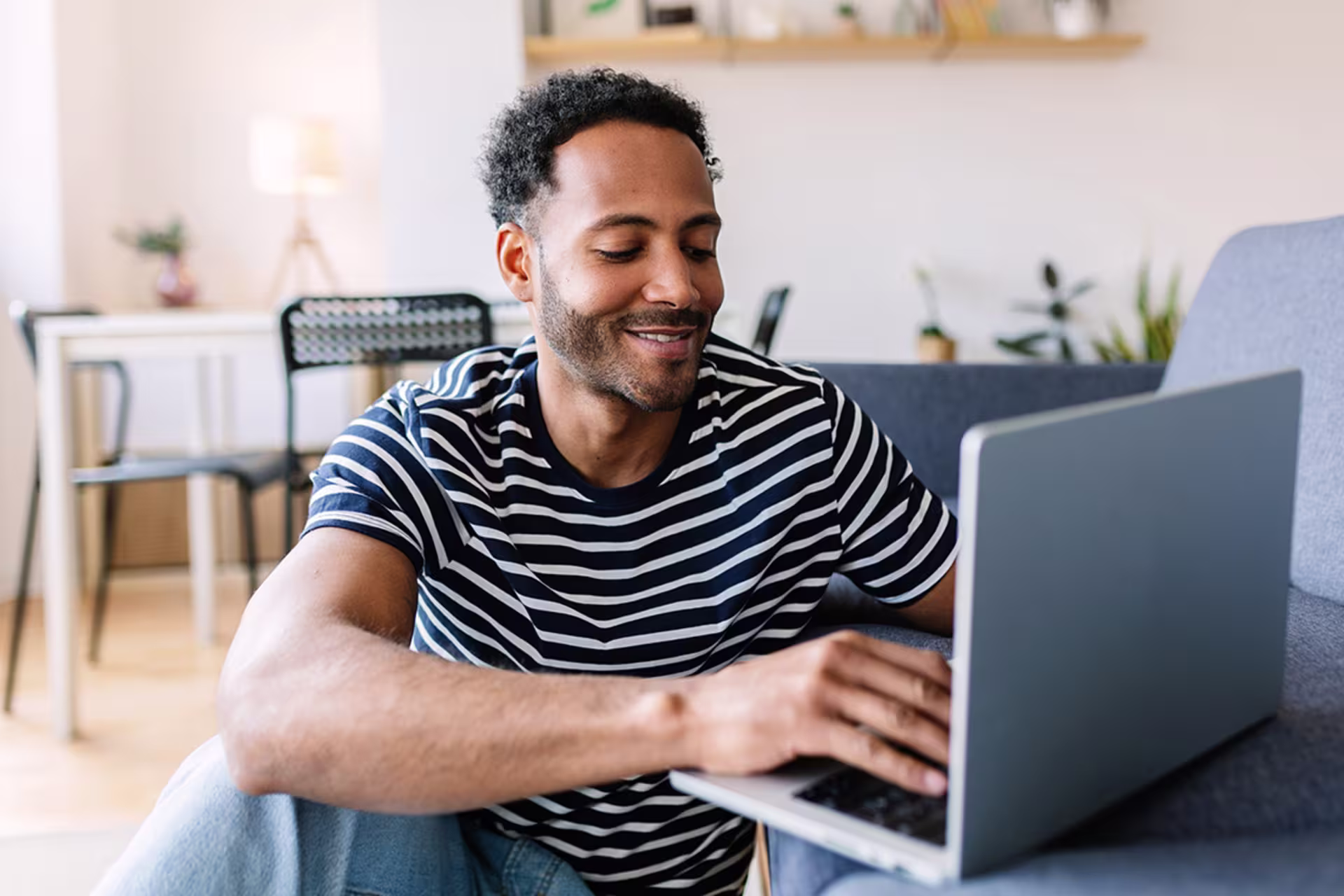 Man smiling while using a laptop on a couch in a bright, modern home office setting.