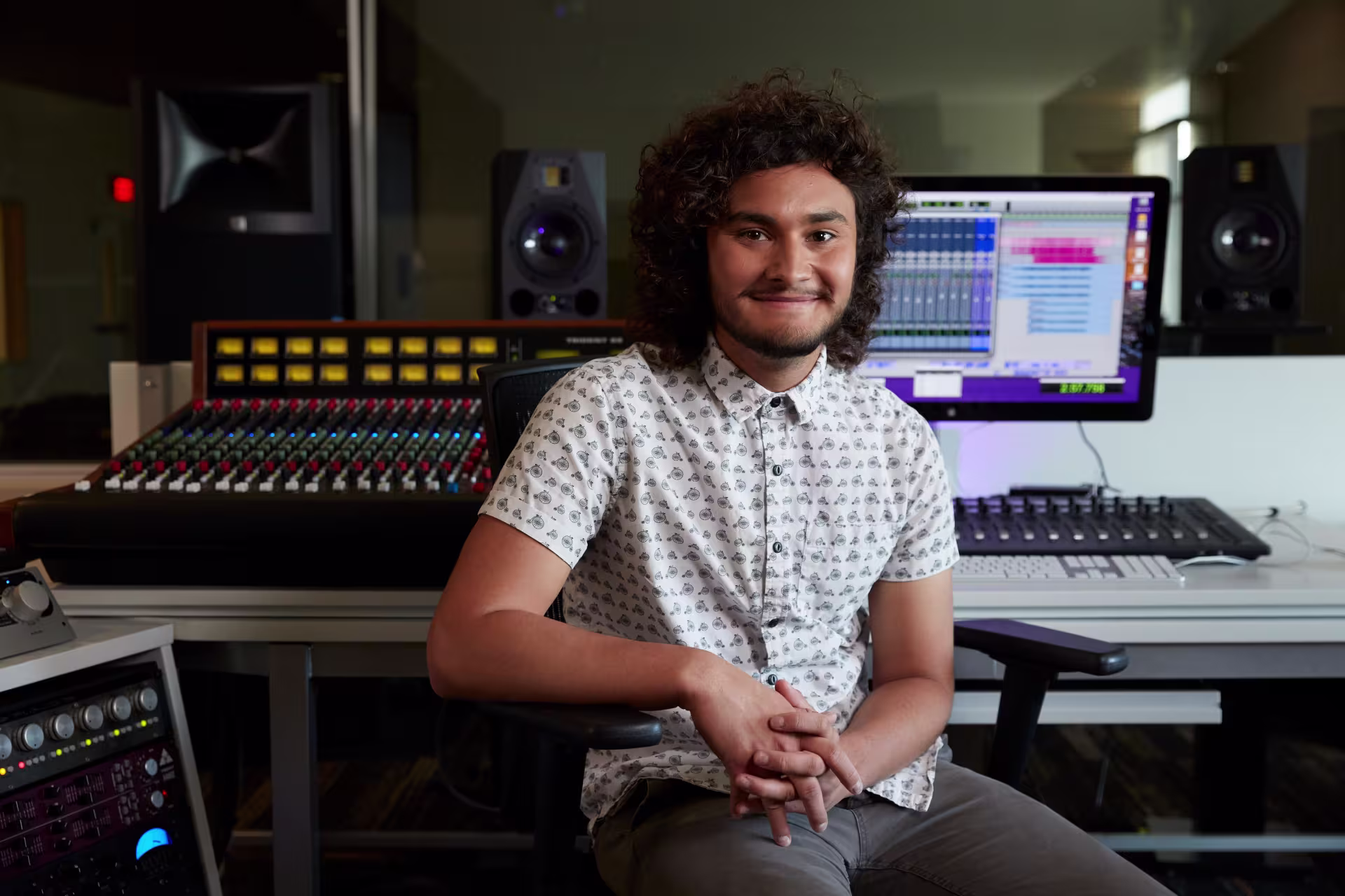Male music production student sitting in a recording studio with a mixing console and digital audio workstation.