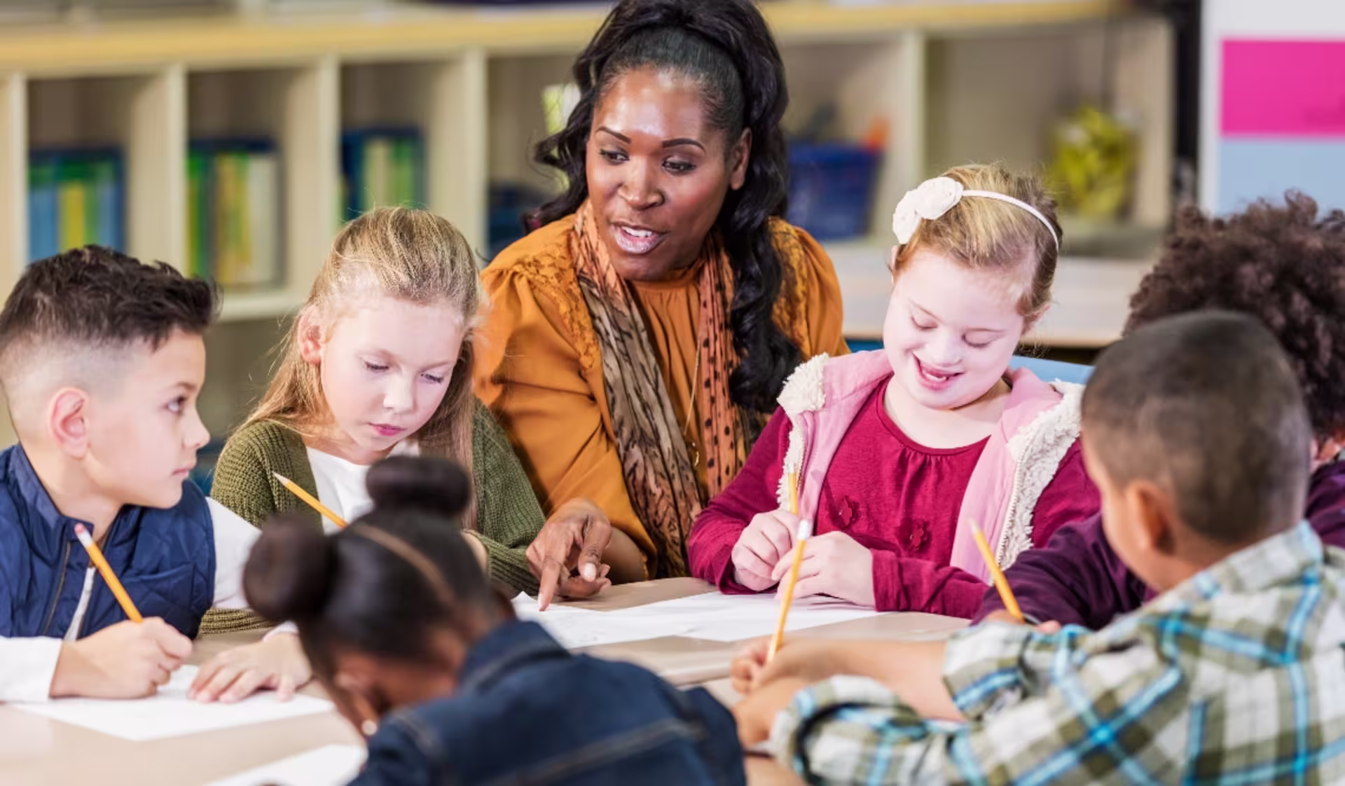 An elementary special education teacher sits with kids at their classroom table while they work on their schoolwork.