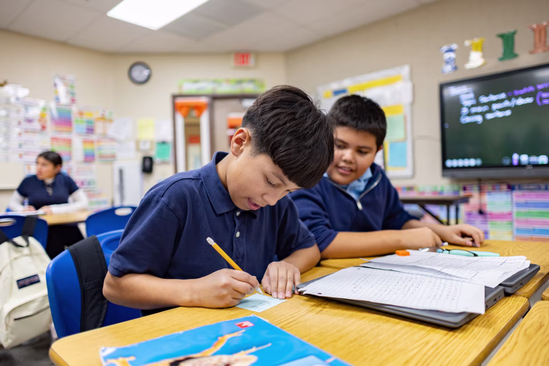 Two male elementary students work together at a classroom desk as one writes with a pencil on a worksheet while the other looks on, with classroom materials and a board in the background.