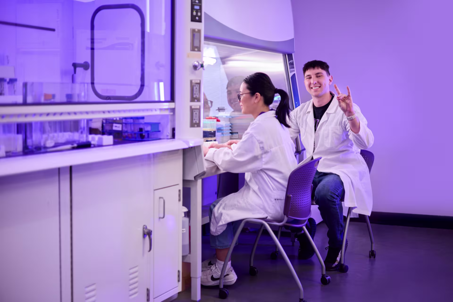 Two students in white lab coats work in a purple-lit biology lab, with one seated at a workstation handling equipment while the other smiles and gives a lopes up sign toward the camera.
