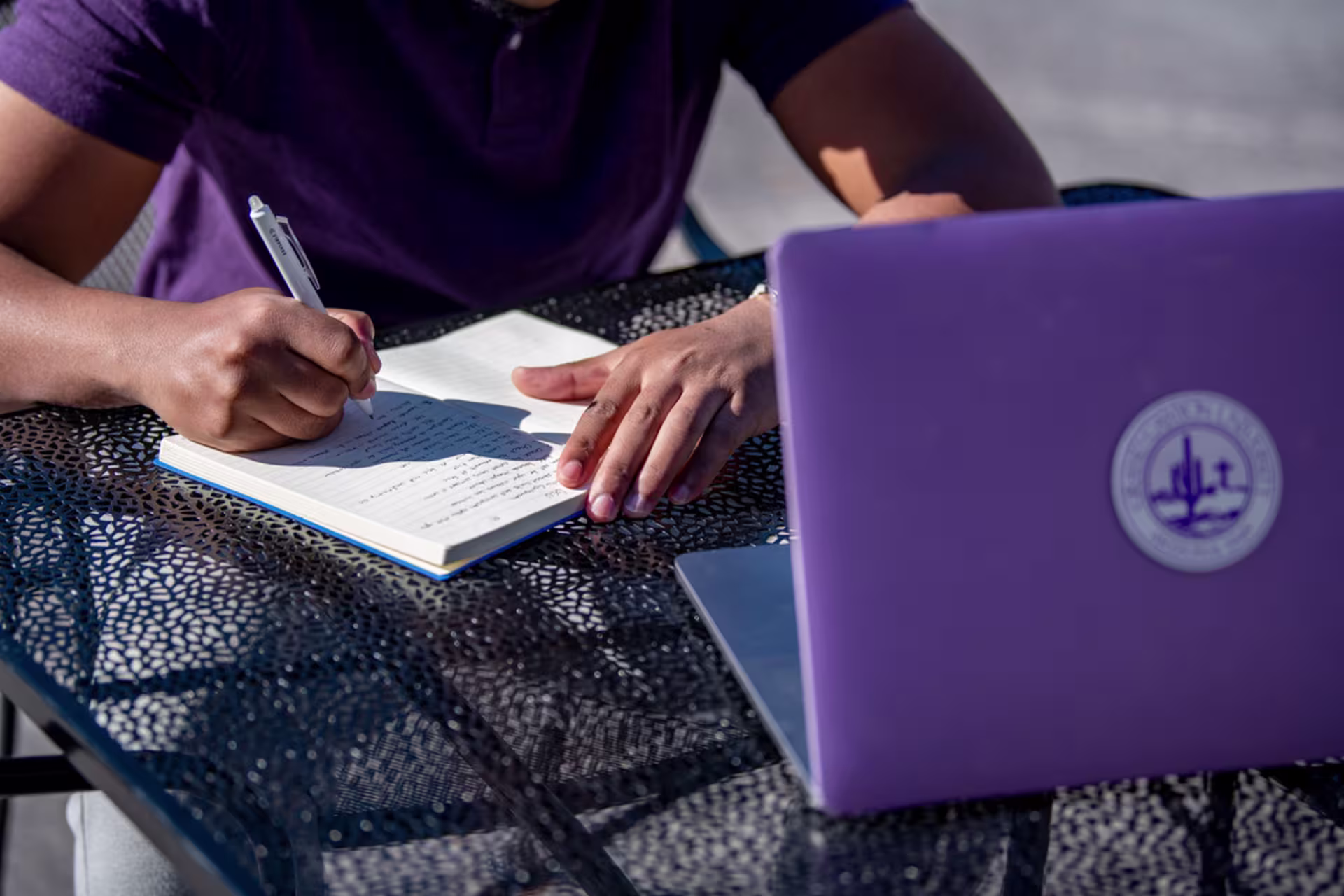 Student taking notes in a notebook beside a laptop outdoors, illustrating writing habits and skills discussed in the professional writing degree at GCU.