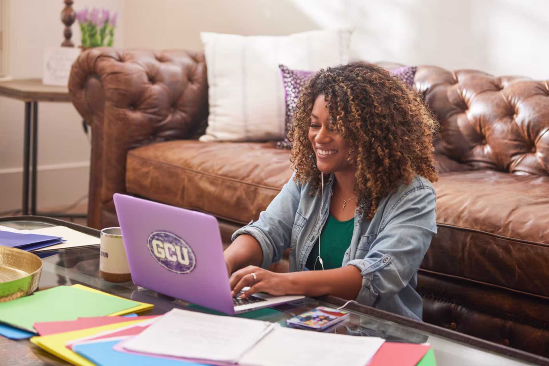 A woman smiles while working on a laptop at a coffee table with notebooks and colorful papers spread out around her.