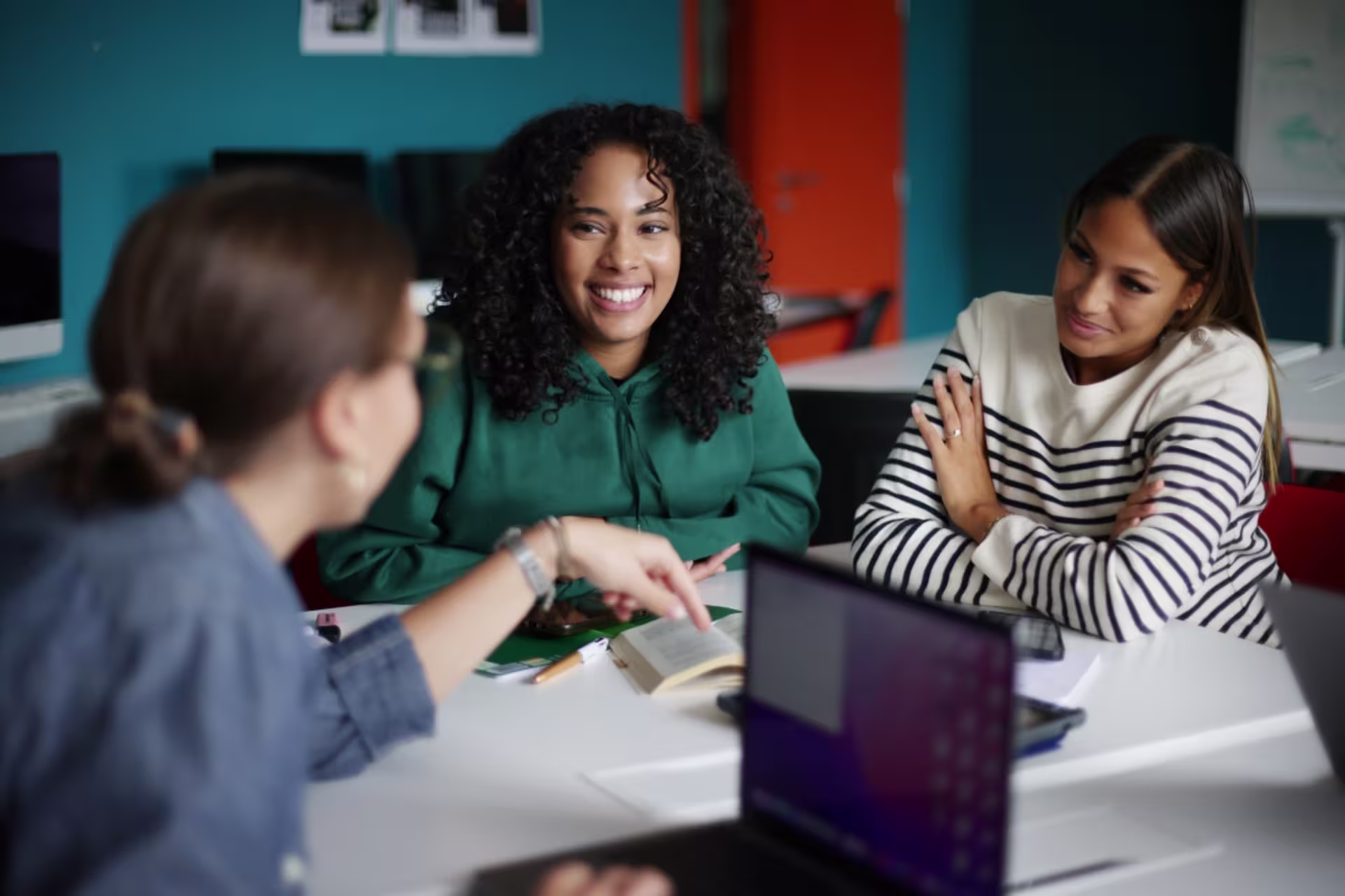 Three women sit together at a table, smiling and talking during a collaborative discussion.