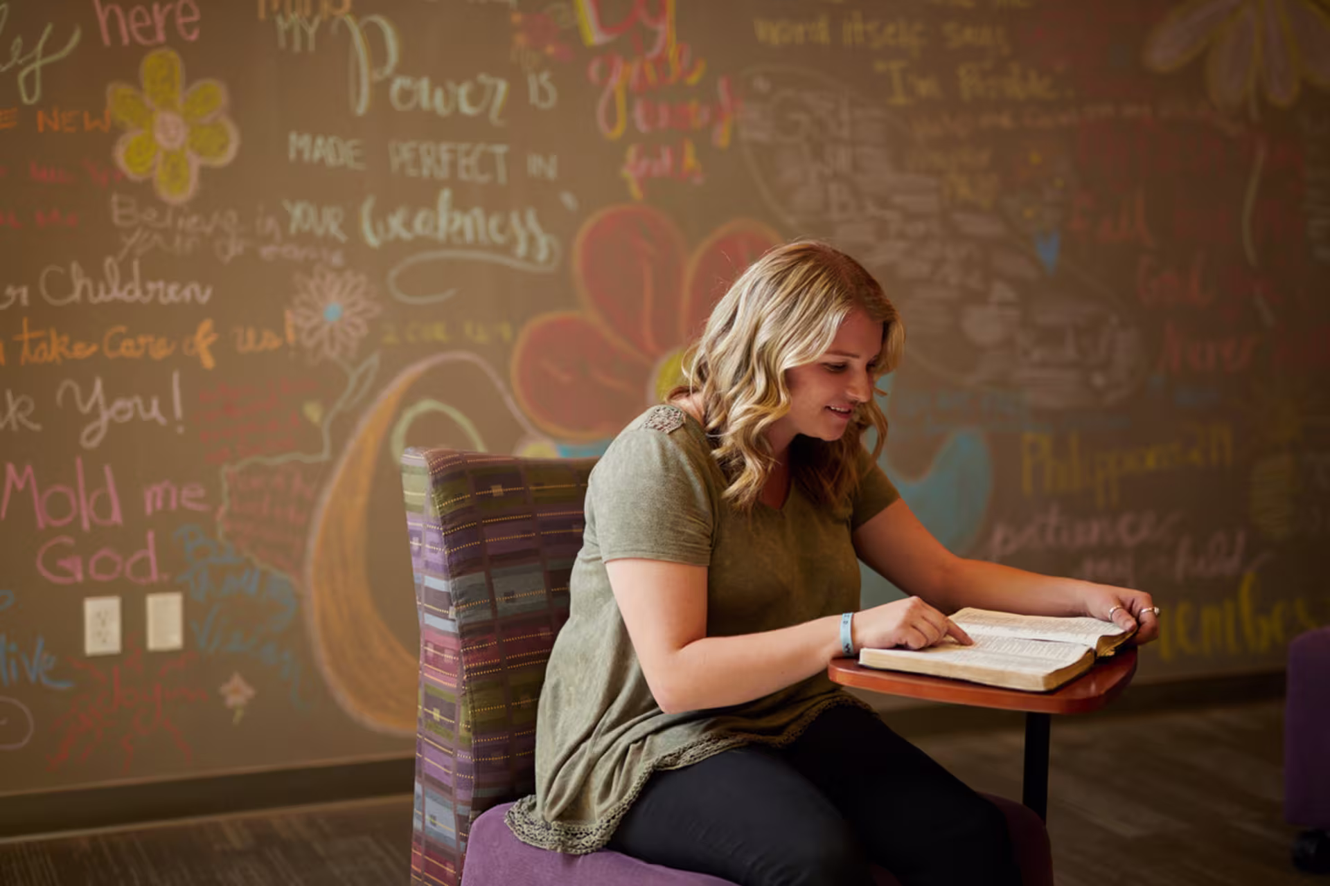 Person seated in a chair reading an open Bible at a small table, with a chalkboard wall behind covered in colorful handwritten words and drawings.