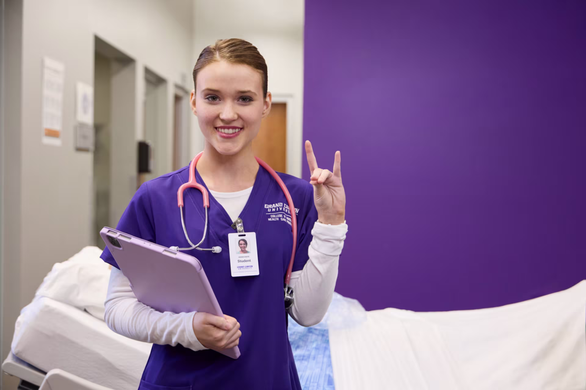A female Boise metro area ABSN student in purple scrubs holds a clipboard while standing in a clinical skills lab setting.