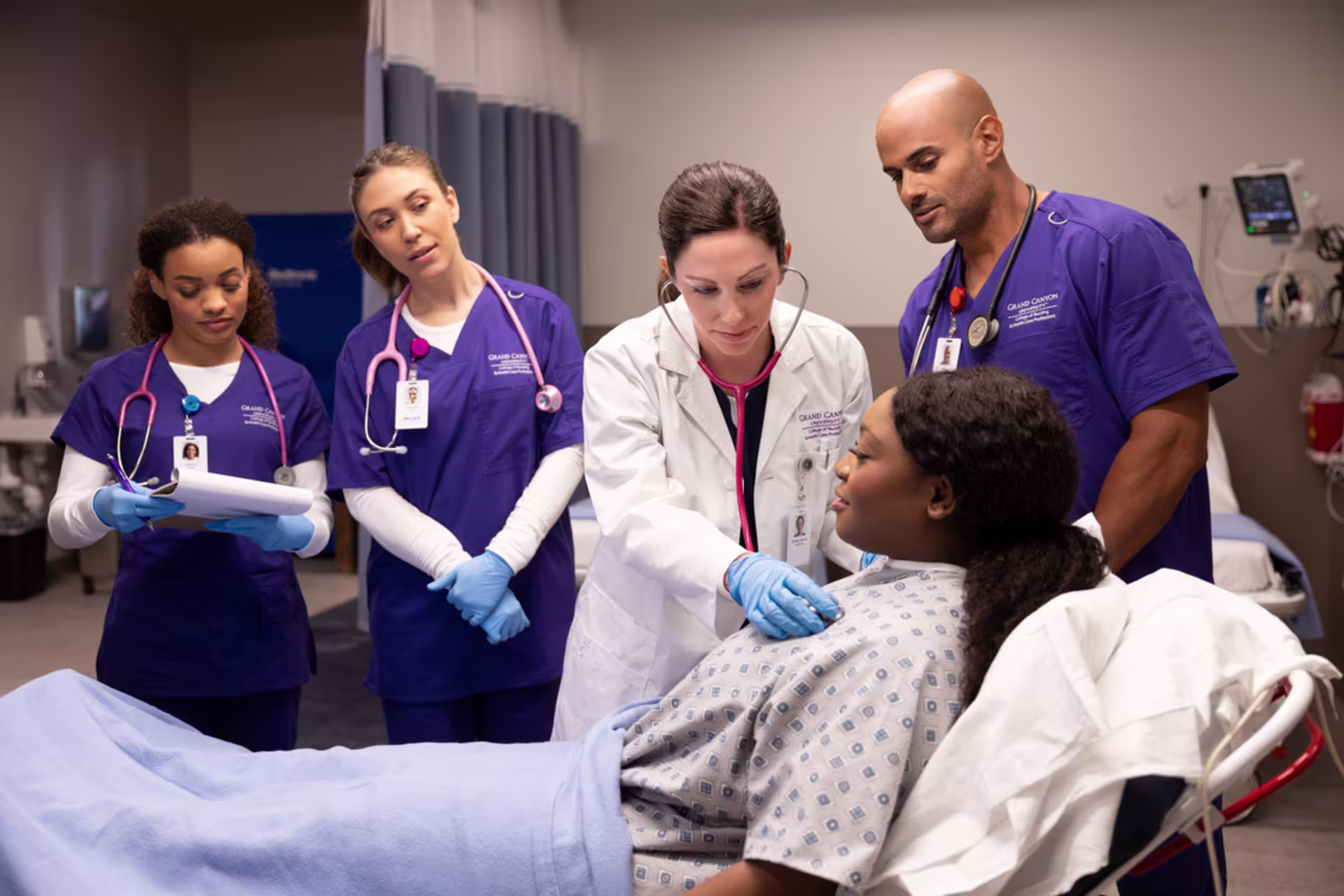 Nursing students in purple scrubs observe an instructor during clinical rotations as she examines a patient in a hospital bed using a stethoscope. One student takes notes while others watch closely.