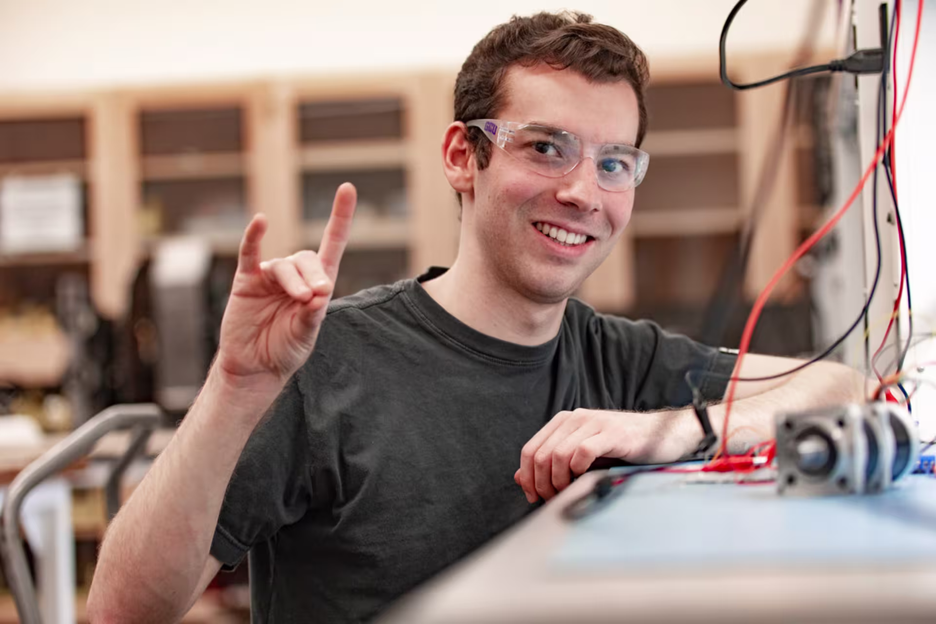 GCU mechanical engineering student in protective eyewear working with wires in a lab while giving the Lopes Up hand signal.