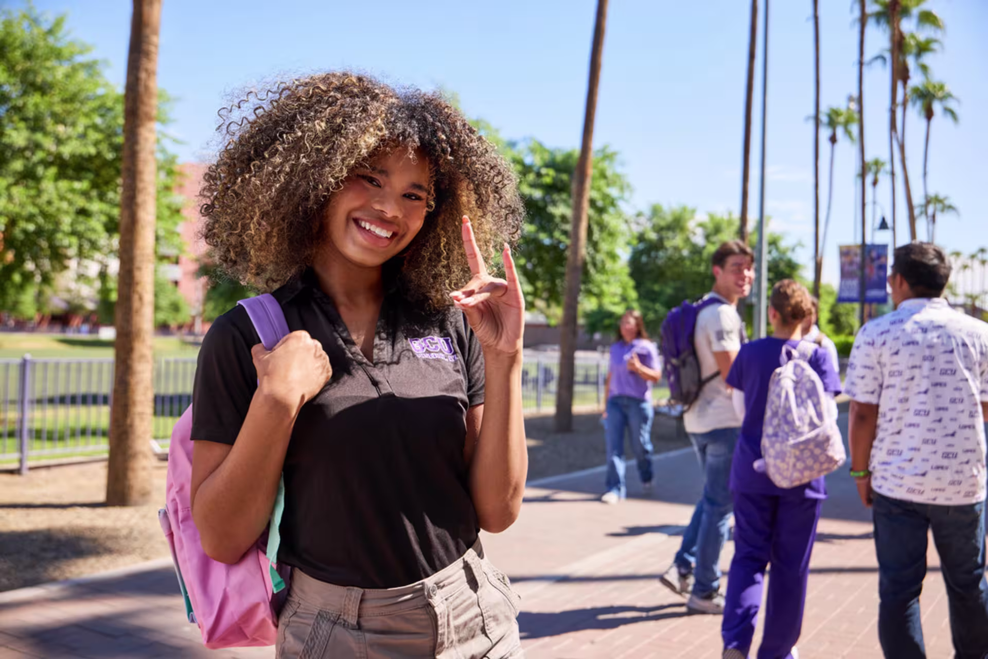 Female GCU bachelor’s student stands on campus with a backpack, smiling and giving the Lopes hand sign with other students in the background.