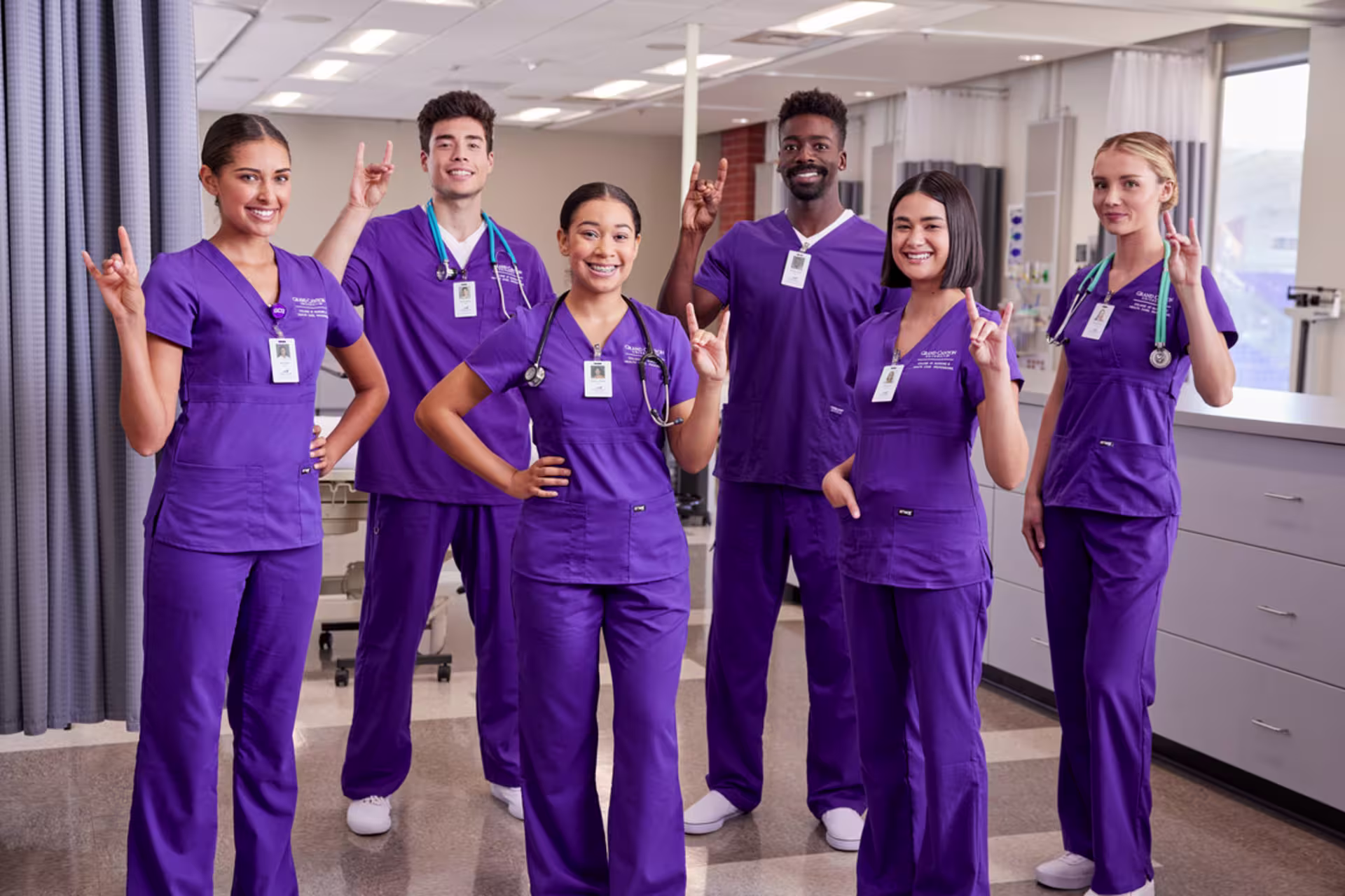 A group of six Florida GCU ABSN students in purple scrubs stand together in a clinical lab setting.