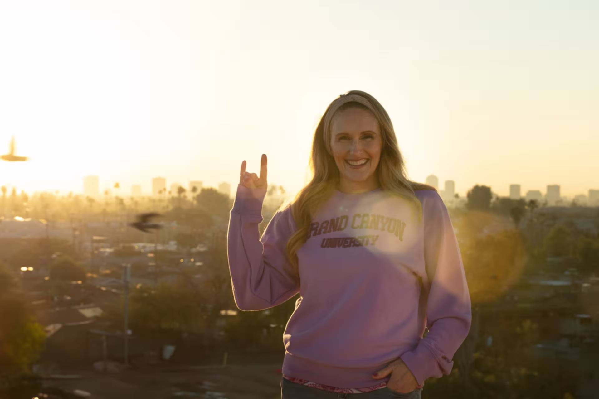 A woman in a GCU sweater shows off the lopes up hand signal, enjoys a sunrise view over a city skyline on a balcony.