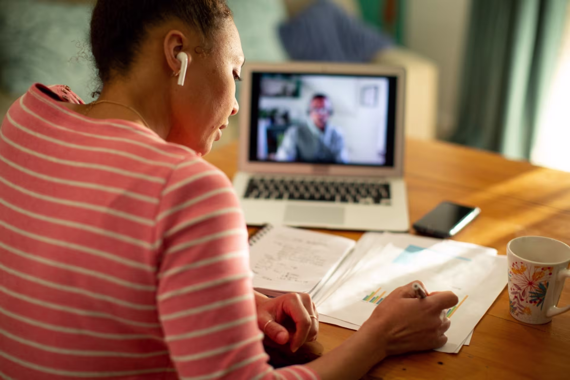 A GCU health informatics student takes notes during an online class on a laptop, wearing earbuds and studying at a desk with a mug and notebook.