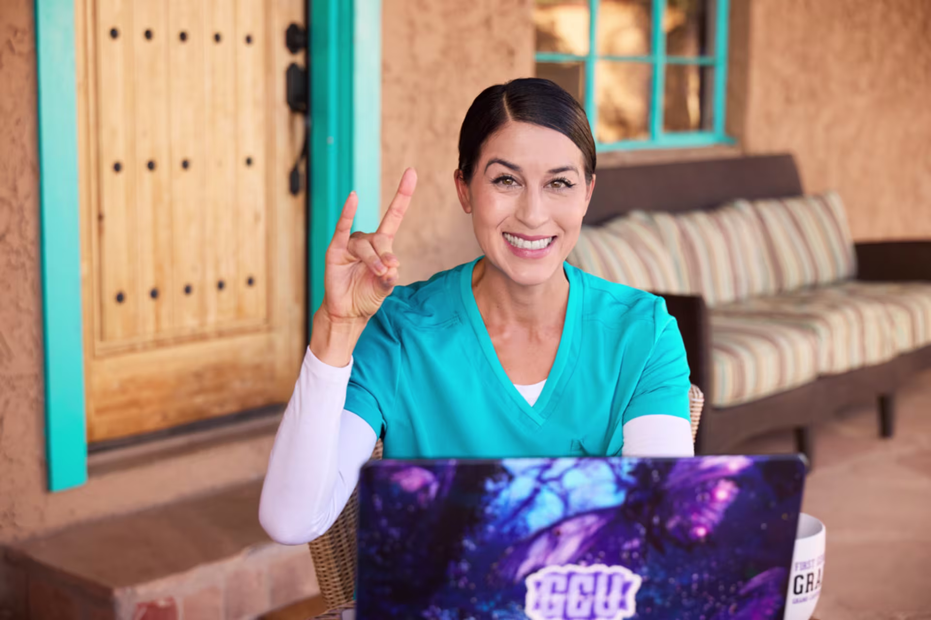 A smiling GCU health informatics student in teal scrubs sits indoors holding a laptop with a colorful abstract screen while raising her hand in greeting.