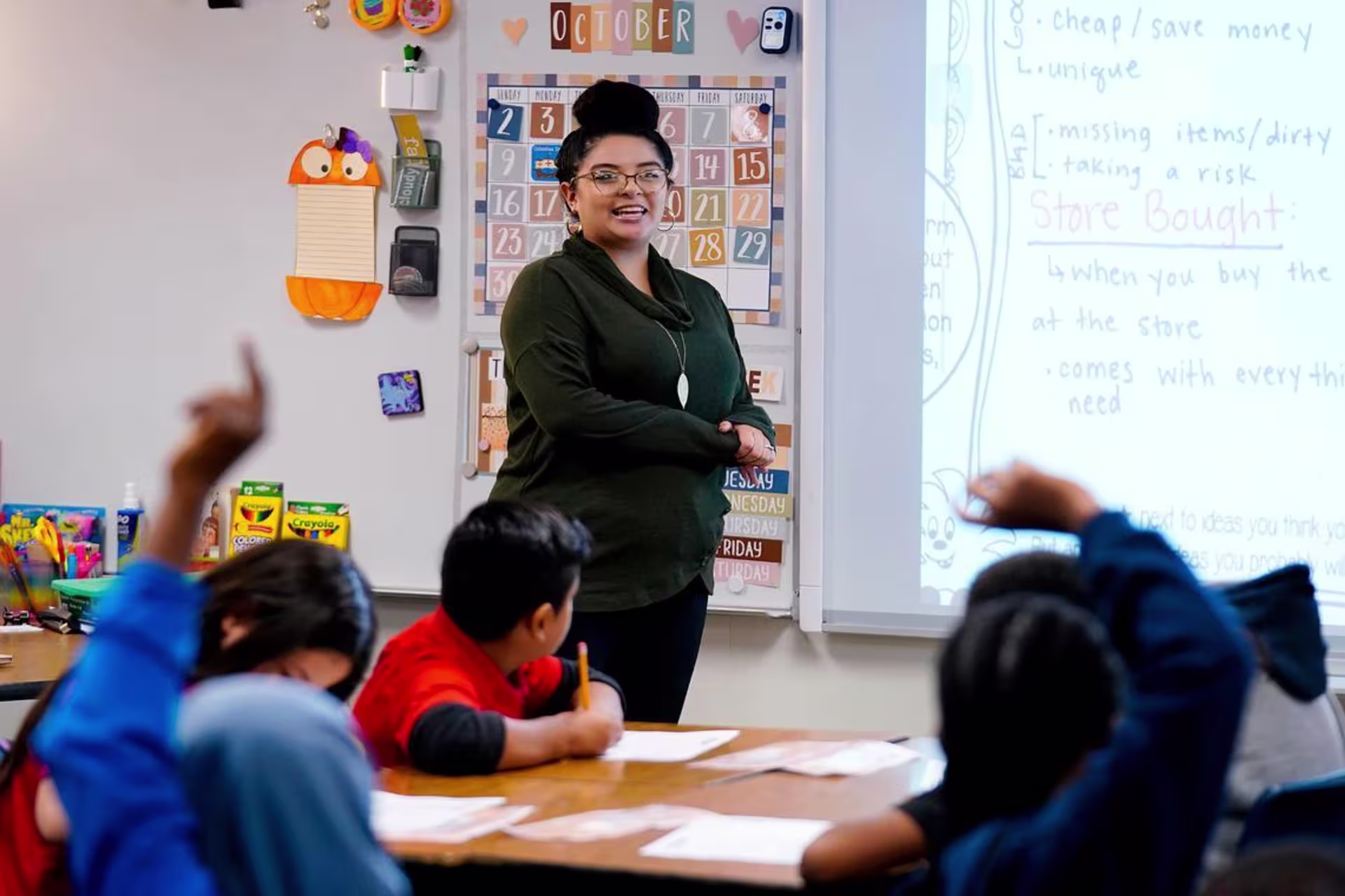 A female teacher stands in front of a class of students taking notes and raising their hands.