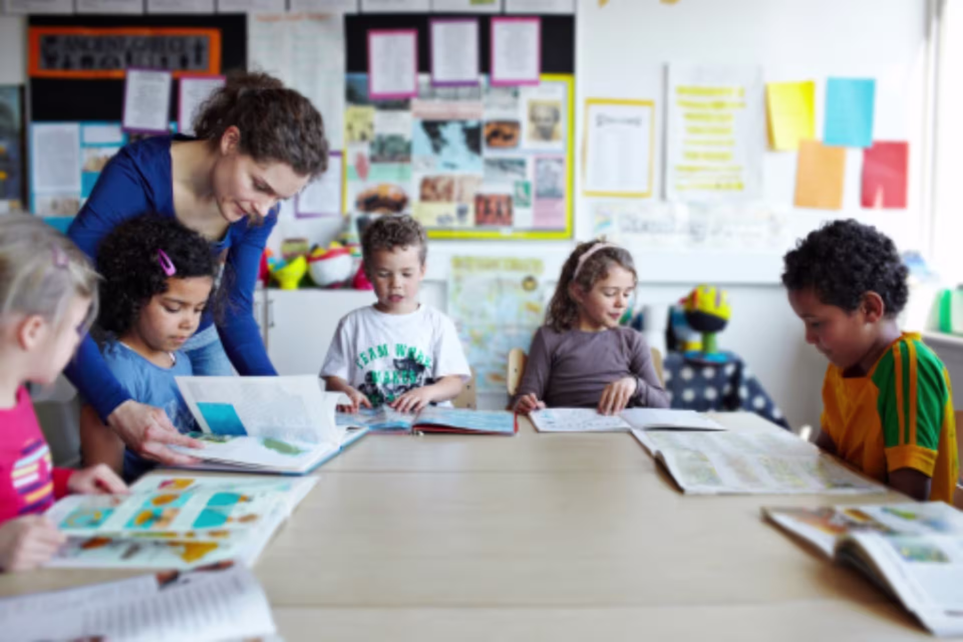 A teacher helps elementary school students read books together at a classroom table.