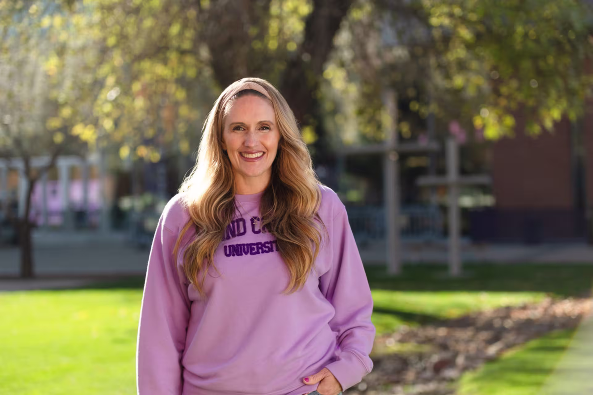 A woman stands outside on a sunny day in a grassy campus setting near crosses, smiling at the camera while wearing a pink sweatshirt that reads Grand Canyon University.