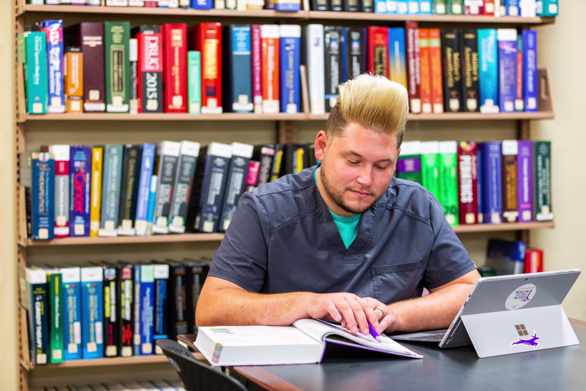 A GCU MSN student sits at a library table studying with an open laptop and several stacked textbooks, focusing on notes in front of him.