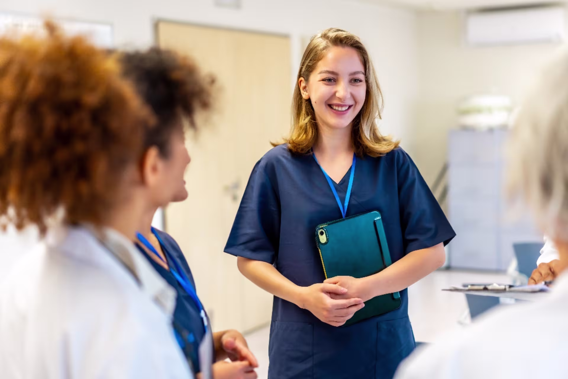 A female MSN student in navy scrubs stands in a clinic, smiling and holding a tablet while speaking with two colleagues.