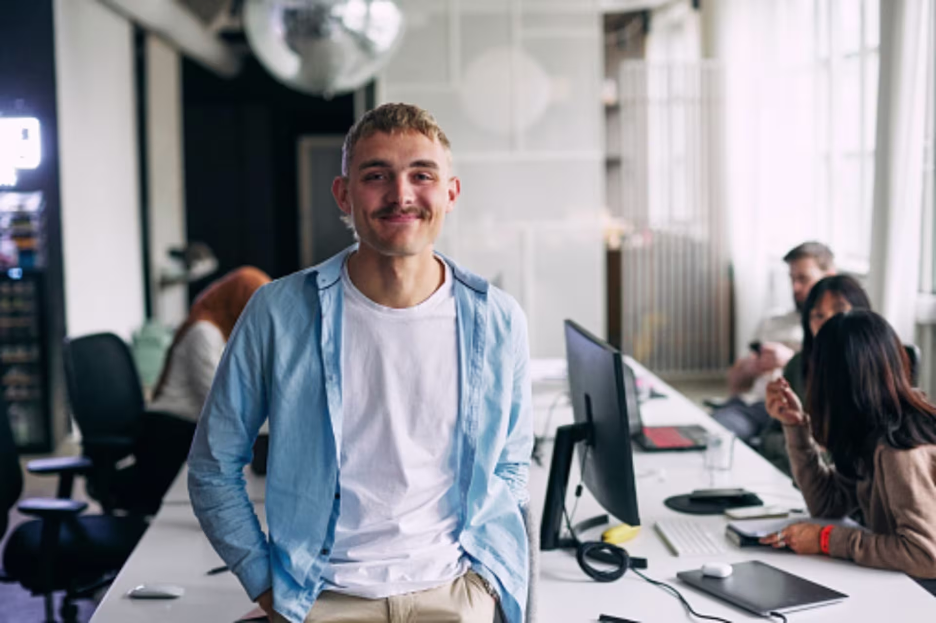 A young male entrepreneur smiles with his hands on his hips in a modern open-concept workspace with colleagues in the background.