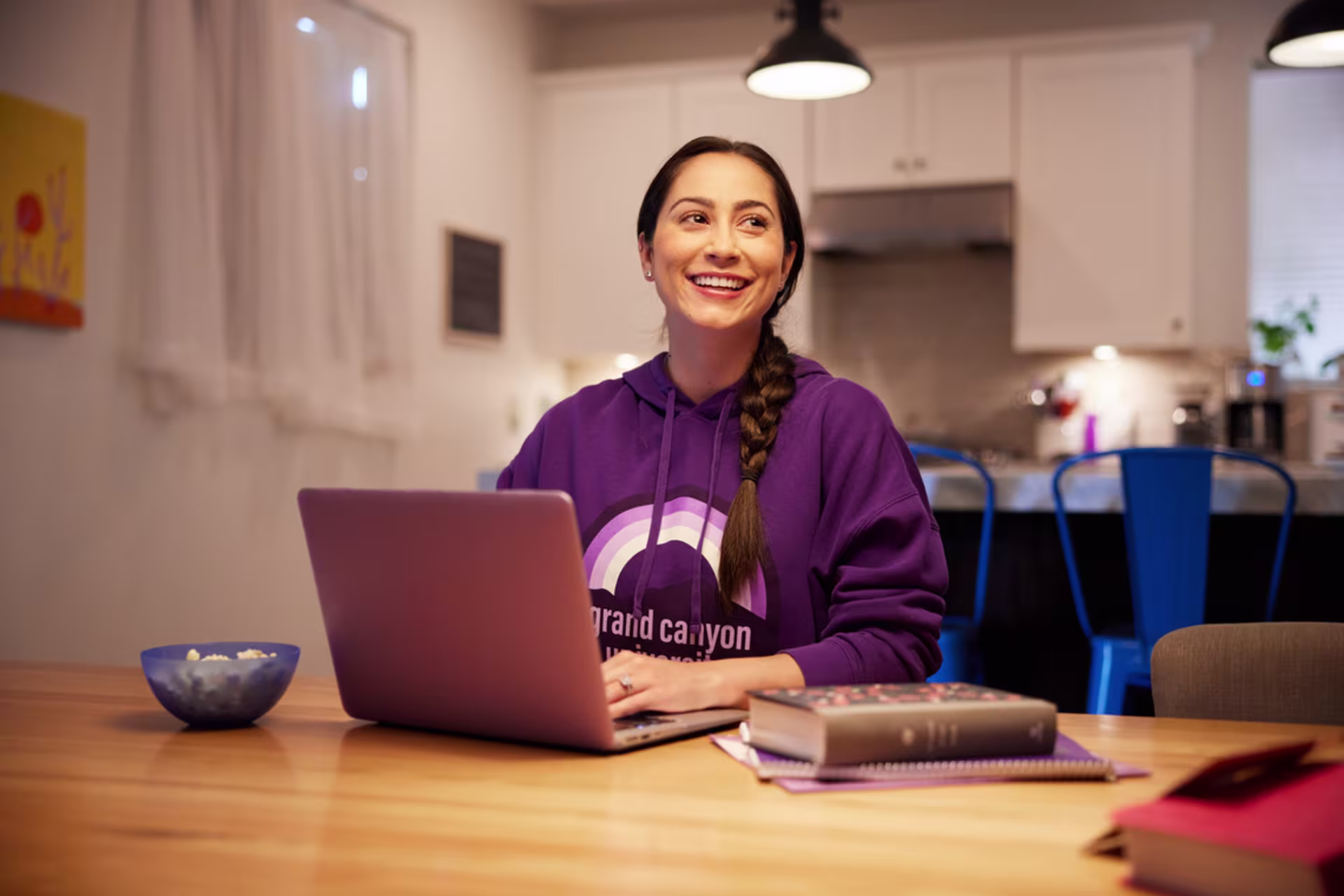 A student studies on a laptop at a dining table with books and notes. Person working at a wooden table with a laptop, notebook and coffee mug in a home workspace, representing independent work and small business planning.