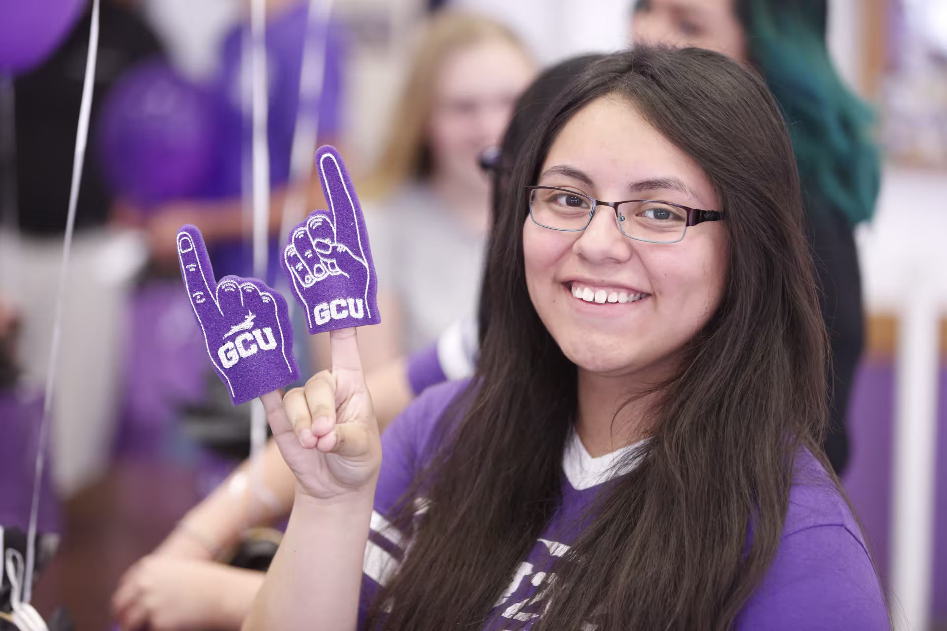 Smiling young woman wearing glasses and a purple shirt holds up two purple GCU foam finger props at a school spirit event with balloons and students in the background.