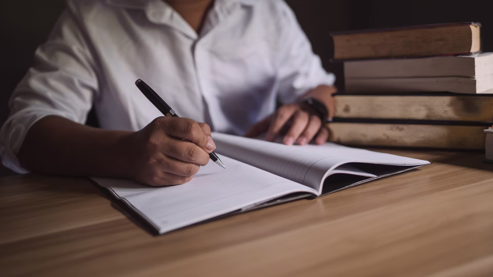 Close-up of a person writing in an open notebook with a pen at a desk, with a stack of books beside them under warm lighting.