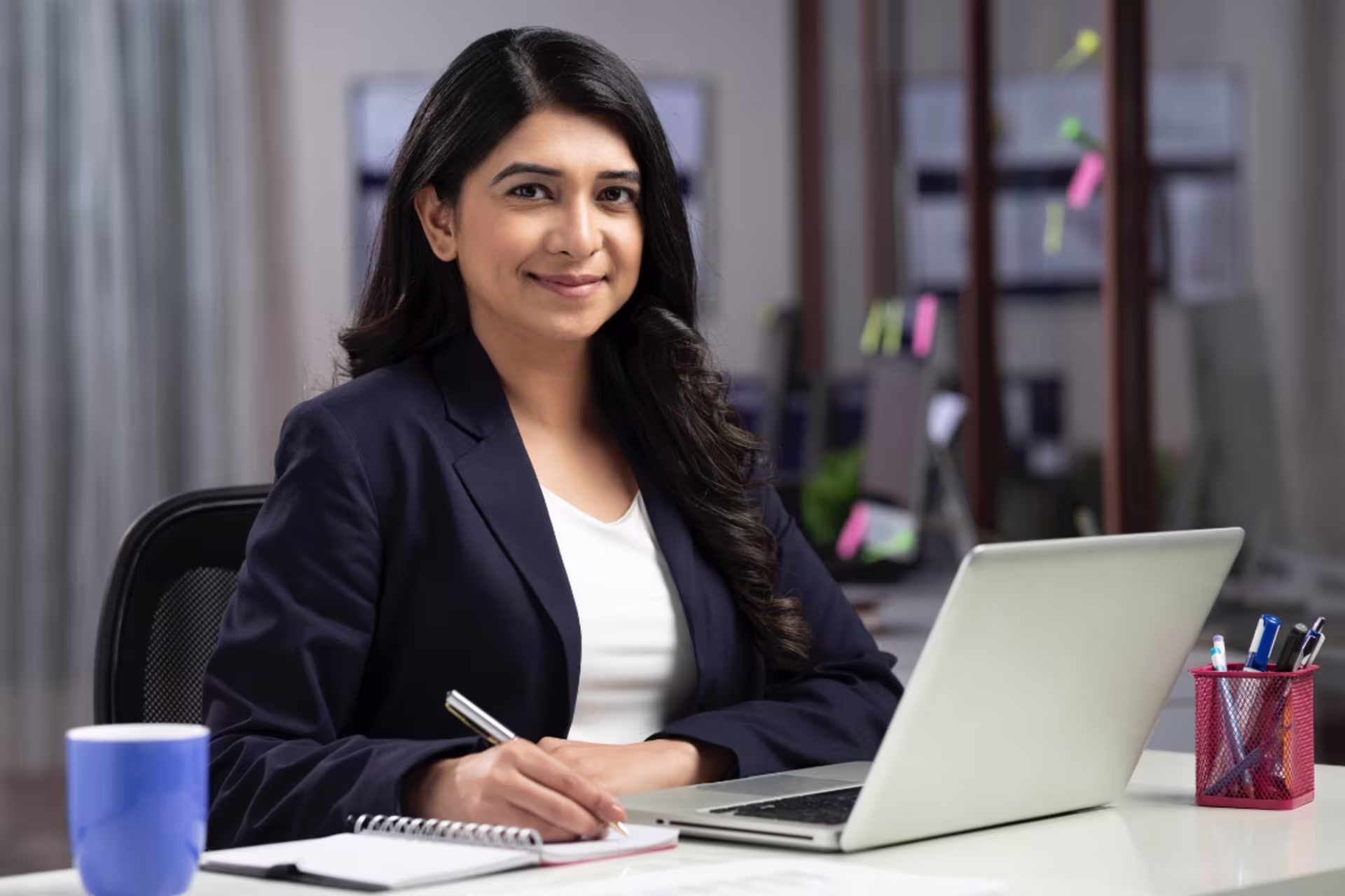 A doctoral business student sits at a desk, smiling while taking notes beside a laptop in a modern office setting.