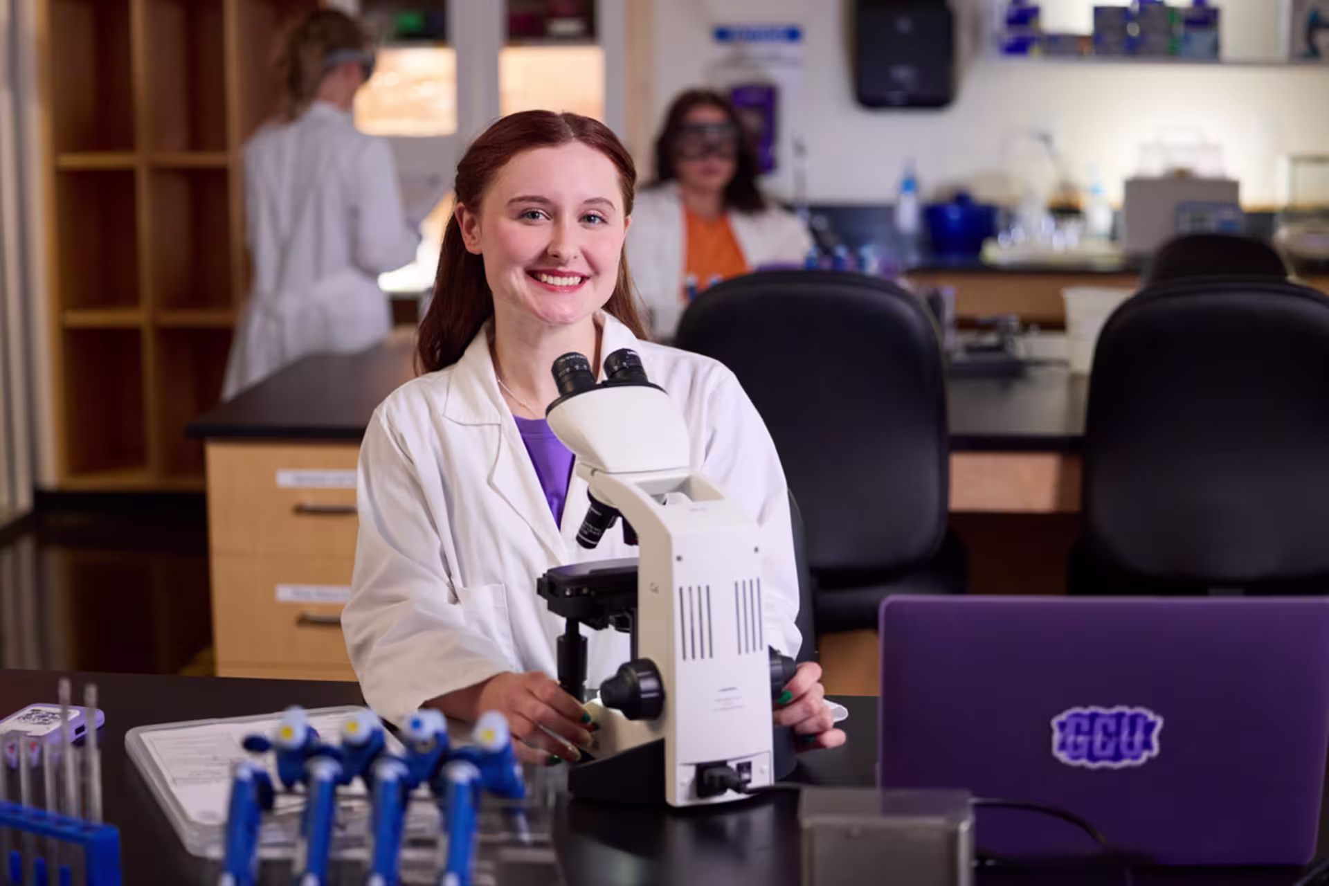 A smiling female pre-PA student uses a microscope in a modern laboratory with lab equipment and colleagues in the background.
