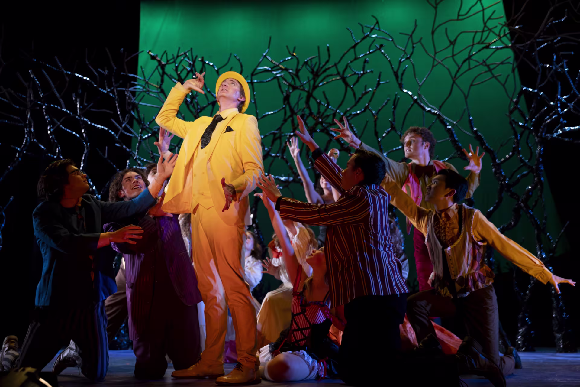 A group of performers on a theater stage surround a central performer in a bright yellow suit, with dramatic lighting and a stylized backdrop of branching shapes.
