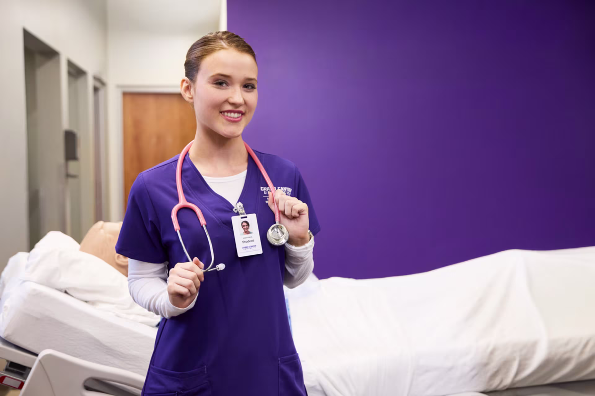 A female nursing student smiles at the camera in a lab setting while standing in front of a sim manikin.