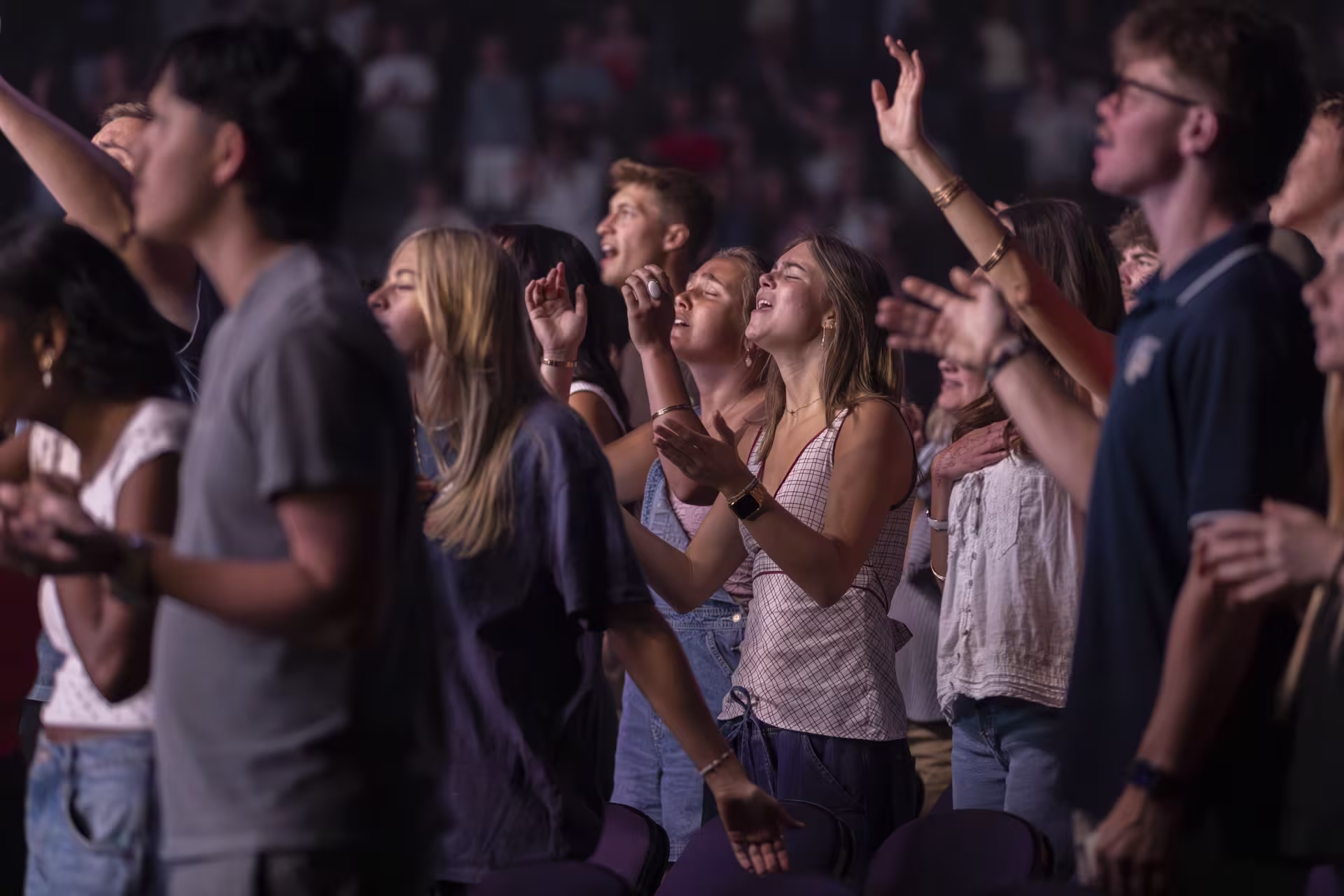 Group of college students standing closely together at a campus worship gathering, raising their hands during a live worship or chapel event.