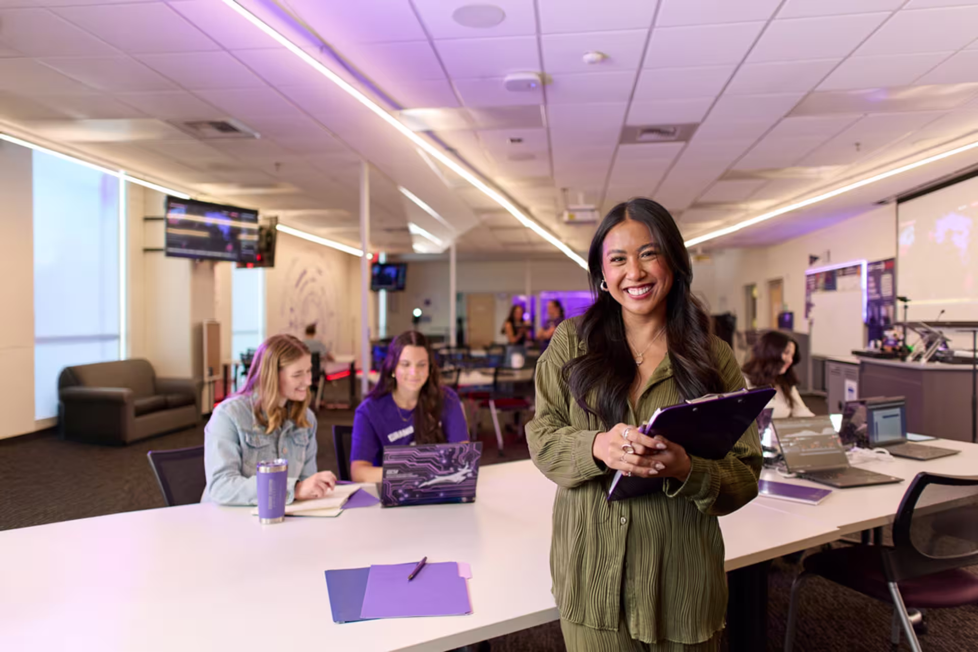 A female GCU MBA student smiles while holding a tablet in a collaborative classroom environment, with classmates working on laptops in the background.
