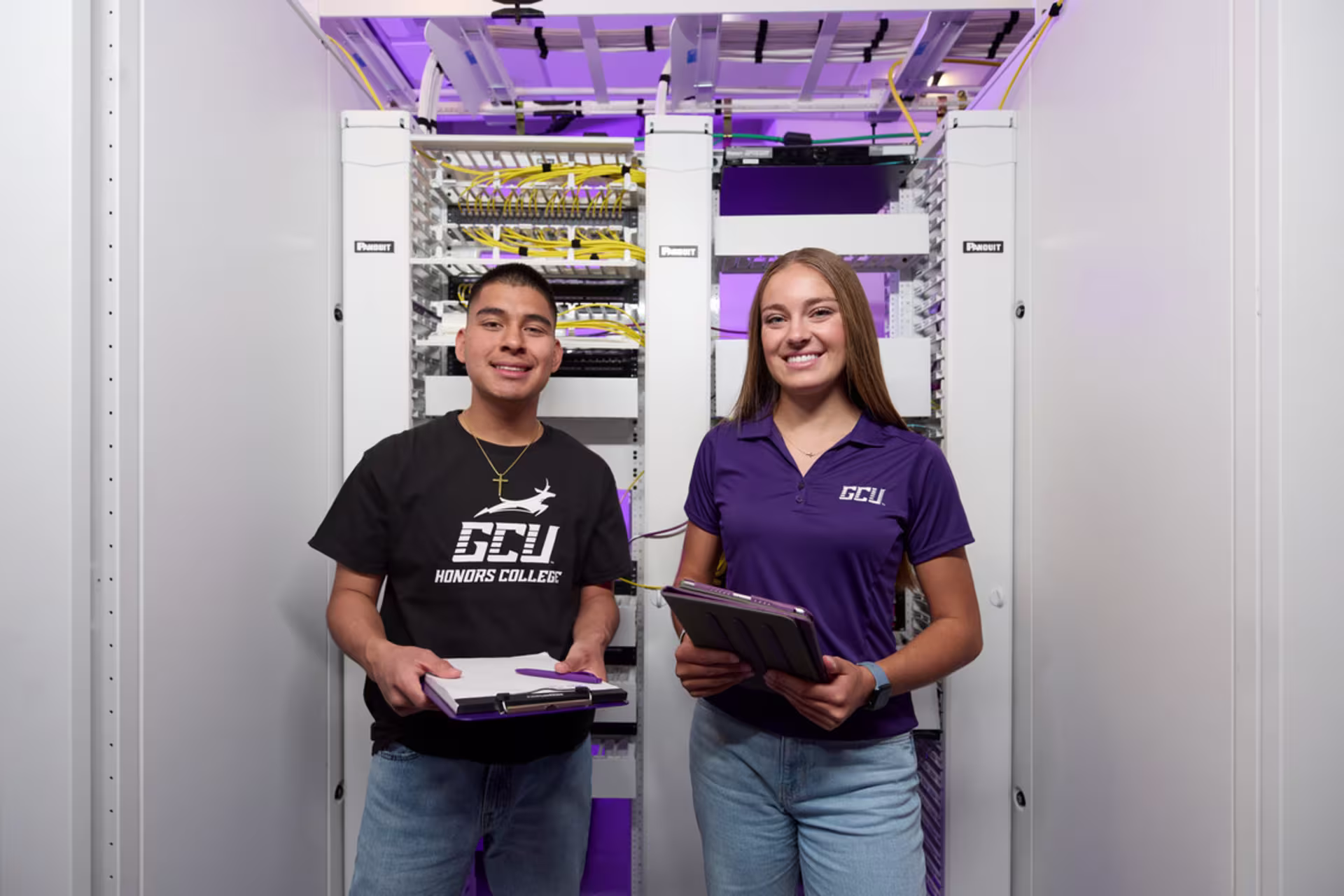 Two GCU cybersecurity students wearing GCU merch hold a clipboard and paperwork while smiling at the camera.
